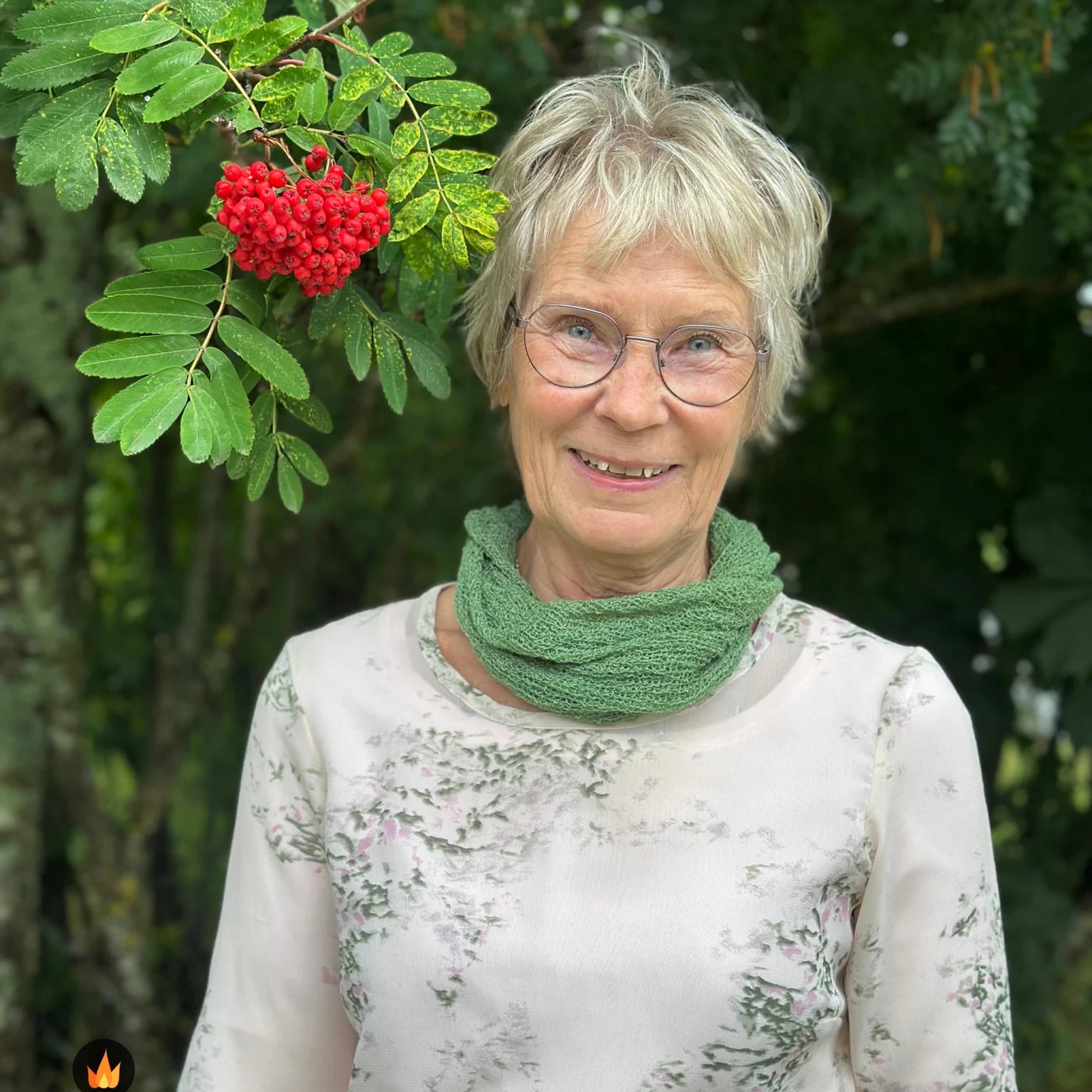 Kvinna bär Eva Tubsjal i färg 034 Ljusgrön, en varm färg i ljus vårgrön, liten finstickad tubsjal buren mjukt runt halsen, fotad utomhus i grönska. Ulla Jacobsson Kläder.