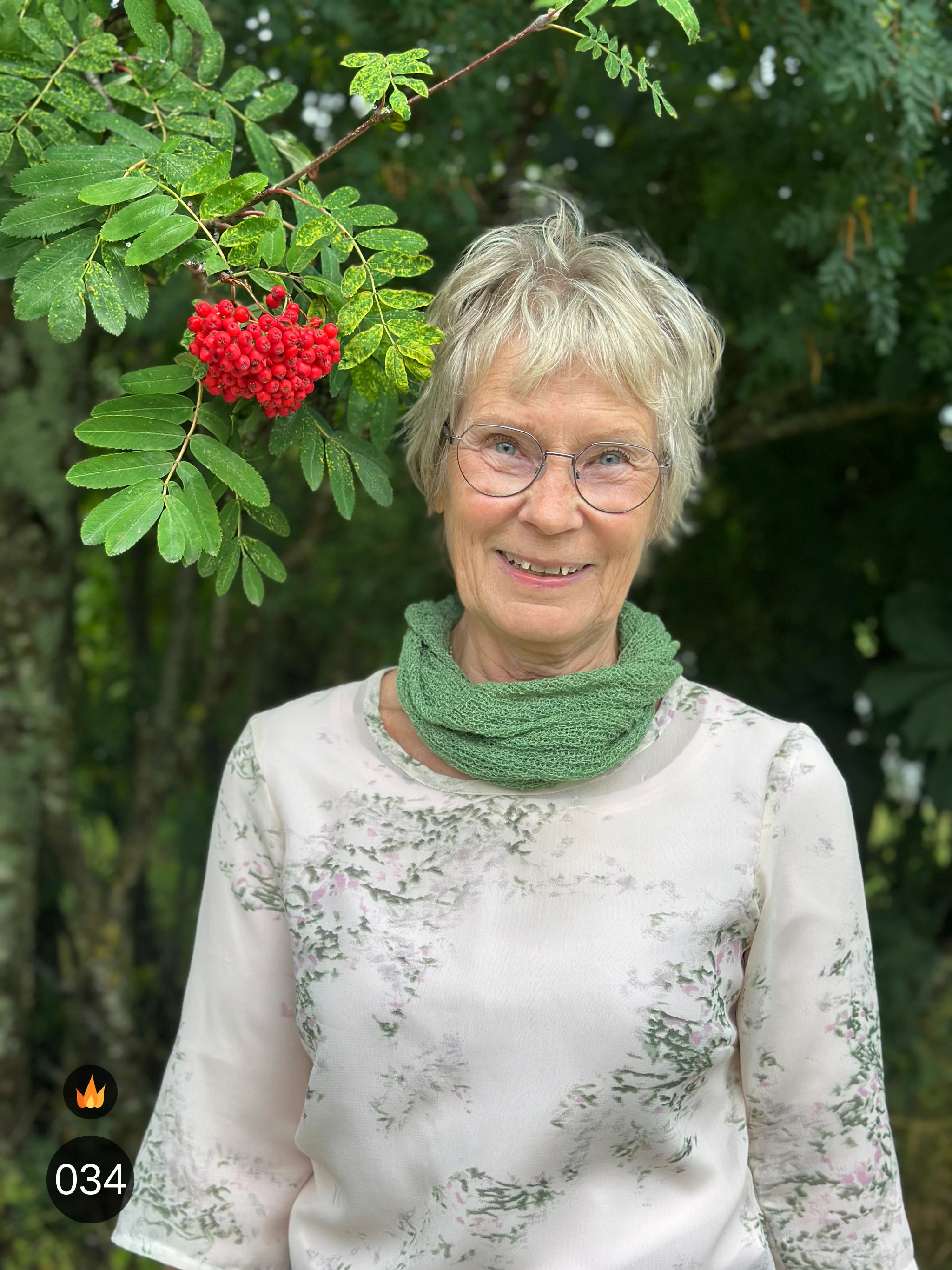 Kvinna bär Eva Tubsjal i färg 034 Ljusgrön, en varm färg i ljus vårgrön, liten finstickad tubsjal buren mjukt runt halsen, fotad utomhus i grönska. Ulla Jacobsson Kläder.