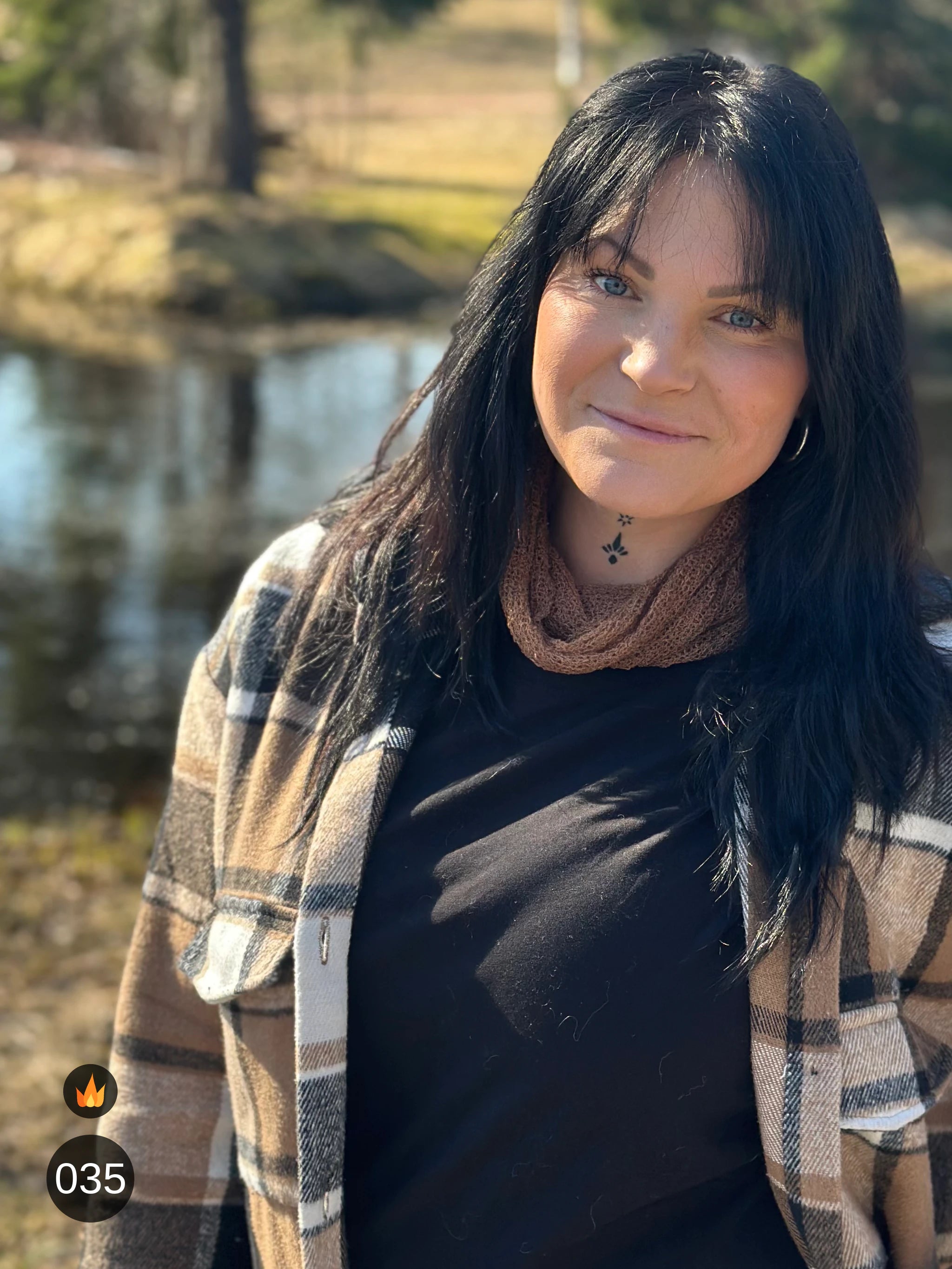 Kvinna bär Eva Tubsjal i färg 035 Bärnsten, en varm färg i varm bärnstensbrun, liten finstickad tubsjal buren mjukt runt halsen, fotad vid vattnet i vårsol. Ulla Jacobsson Kläder.