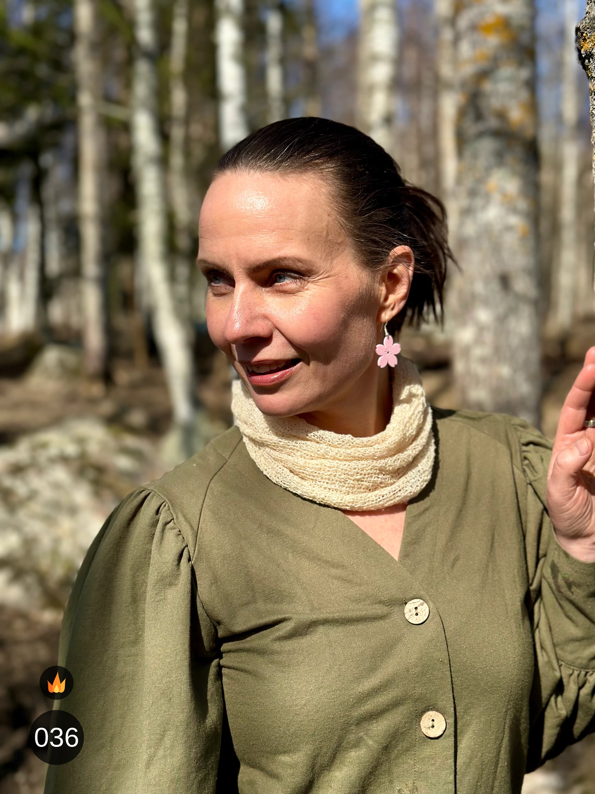 Kvinna bär Eva Tubsjal i färg 036 Ljus sand, en varm färg i ljus sandbeige med champagneton, liten finstickad tubsjal buren mjukt runt halsen, fotad bland björkstammar en vårdag. Ulla Jacobsson Kläder.