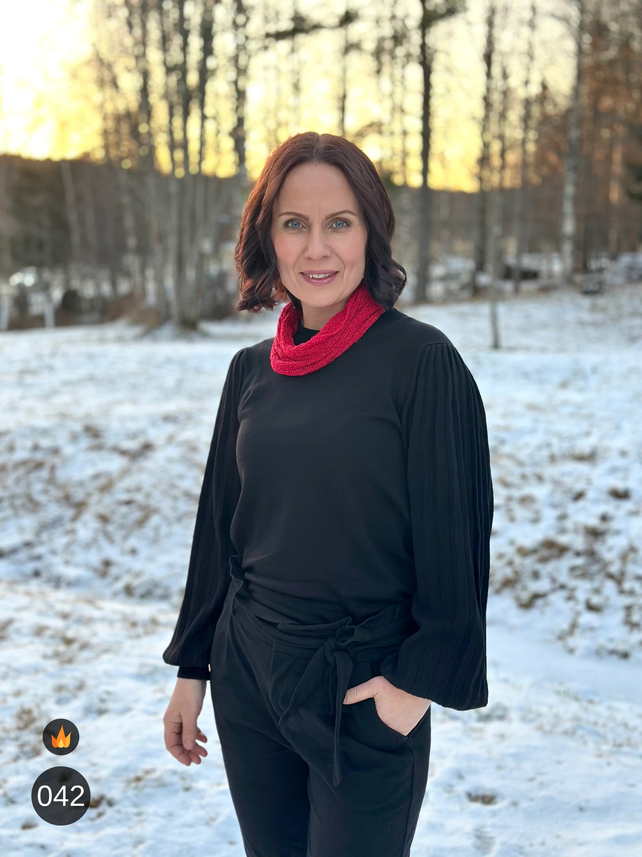 Kvinna bär Eva Tubsjal i färg 042 Röd, en varm färg i klar röd, liten finstickad tubsjal buren mjukt runt halsen, fotad i snöig björkskog i lågt kvällsljus. Ulla Jacobsson Kläder.