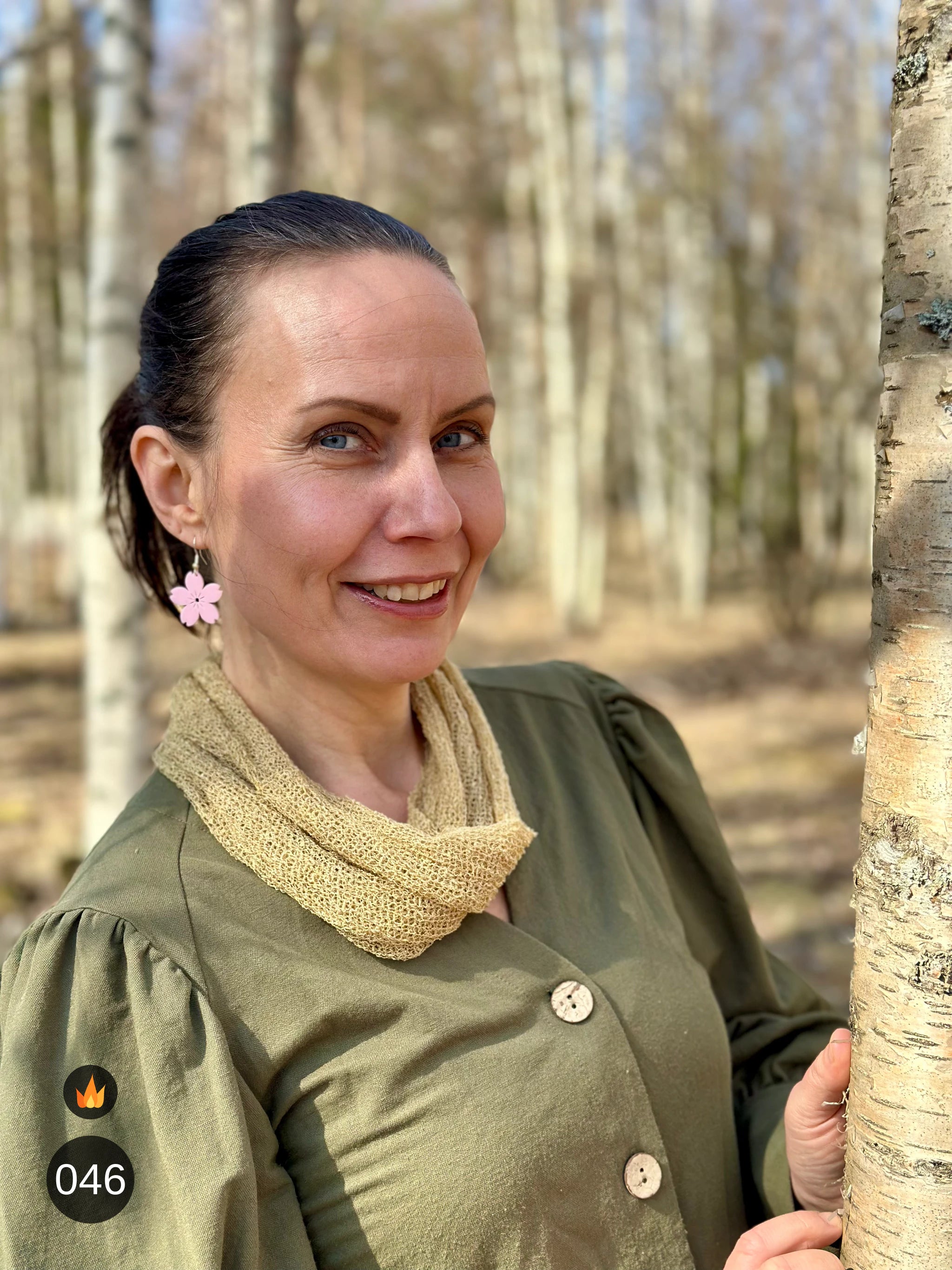 Kvinna bär Eva Tubsjal i färg 046 Havre, en varm färg i ljus havregul, liten finstickad tubsjal buren mjukt runt halsen, fotad vid en björkstam en vårdag. Ulla Jacobsson Kläder.