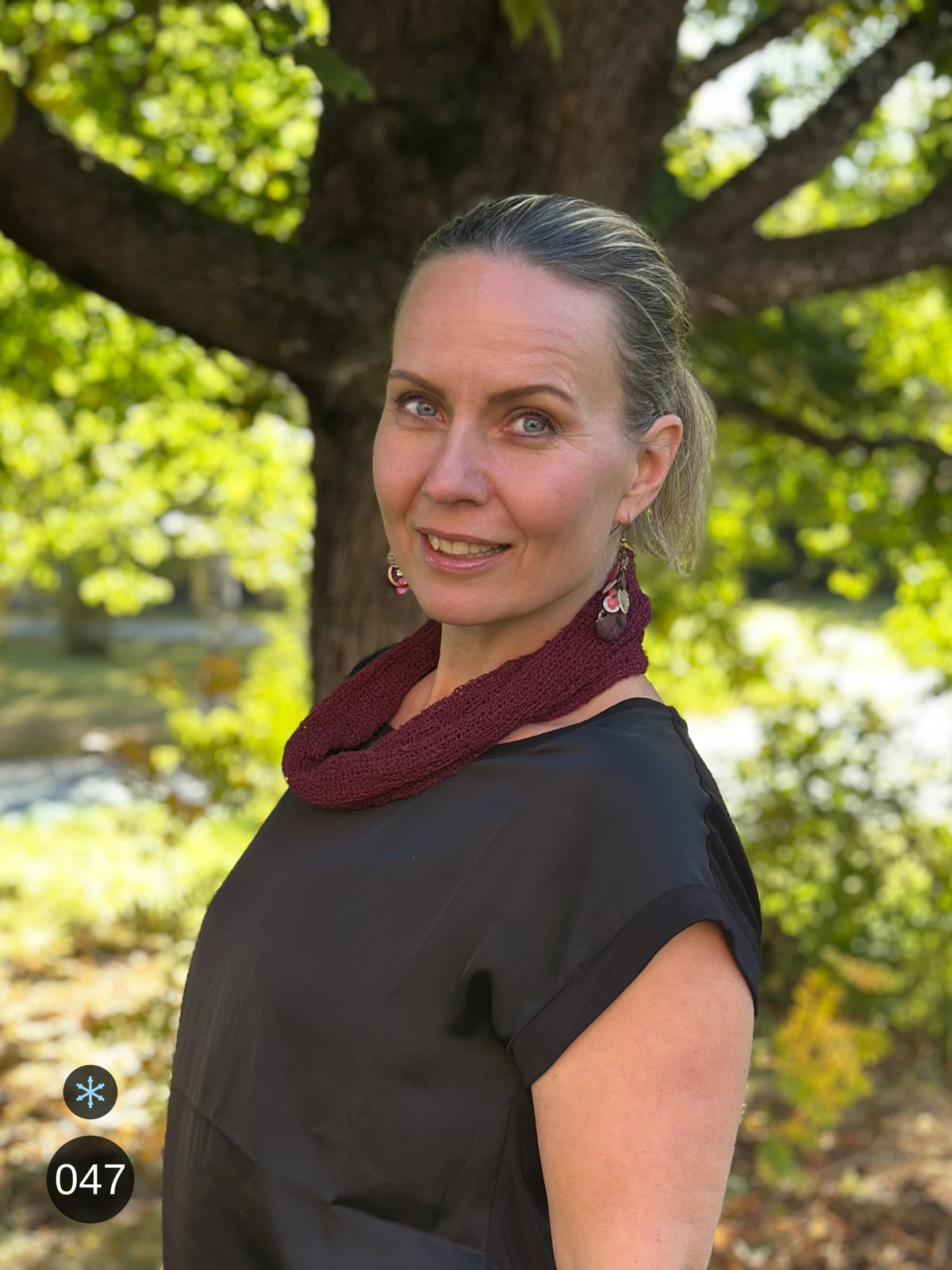 Kvinna bär Eva Tubsjal i färg 047 Bourdeaux, en kall färg i djup bourdeauxröd med blå underton, liten finstickad tubsjal buren som krage, fotad i höstsol under en lönn. Ulla Jacobsson Kläder.