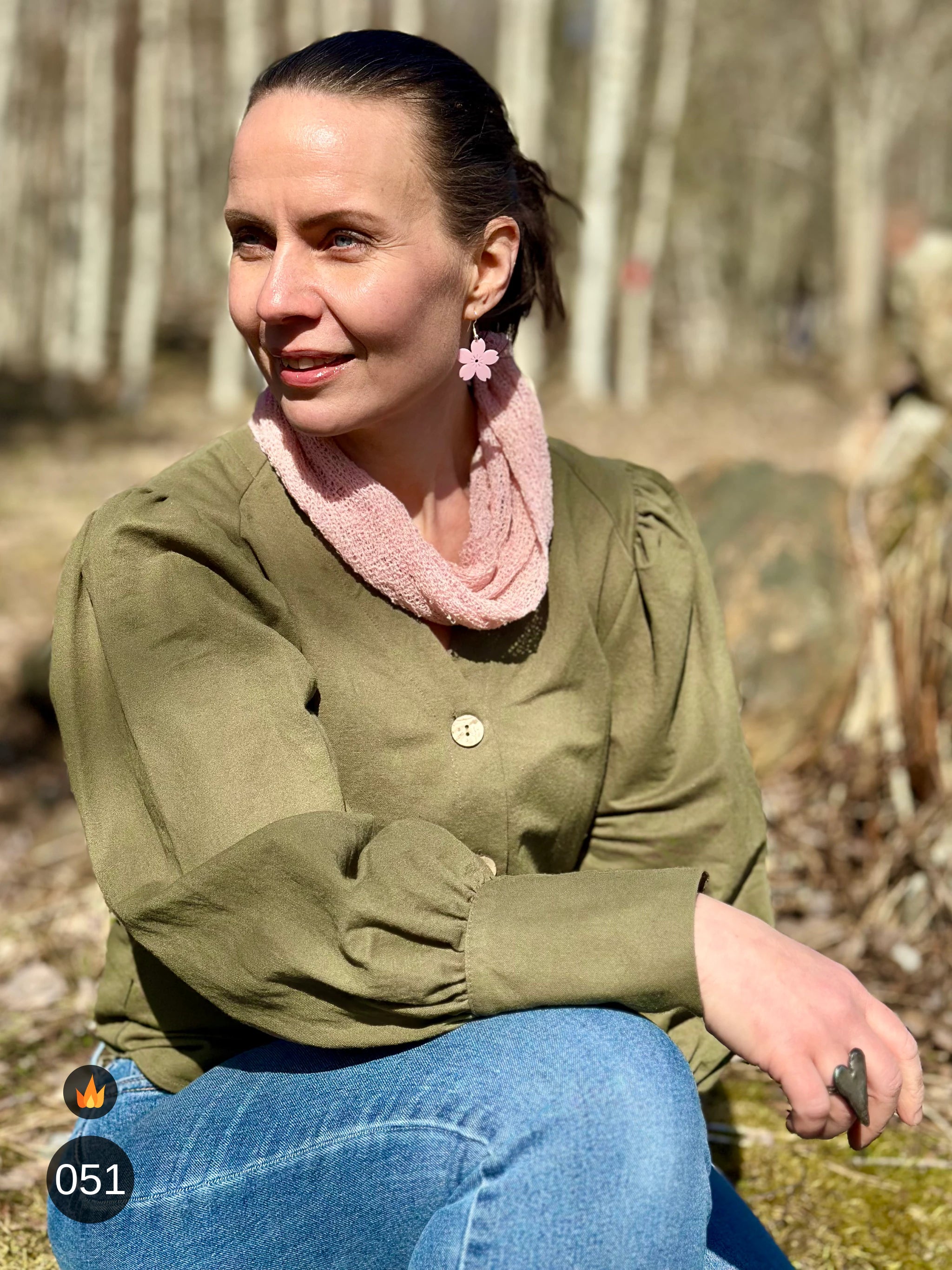 Kvinna bär Eva Tubsjal i färg 051 Puderrosa, en varm färg i mjuk puderrosa, liten finstickad tubsjal buren mjukt runt halsen, fotad sittande på en sten en vårdag. Ulla Jacobsson Kläder.
