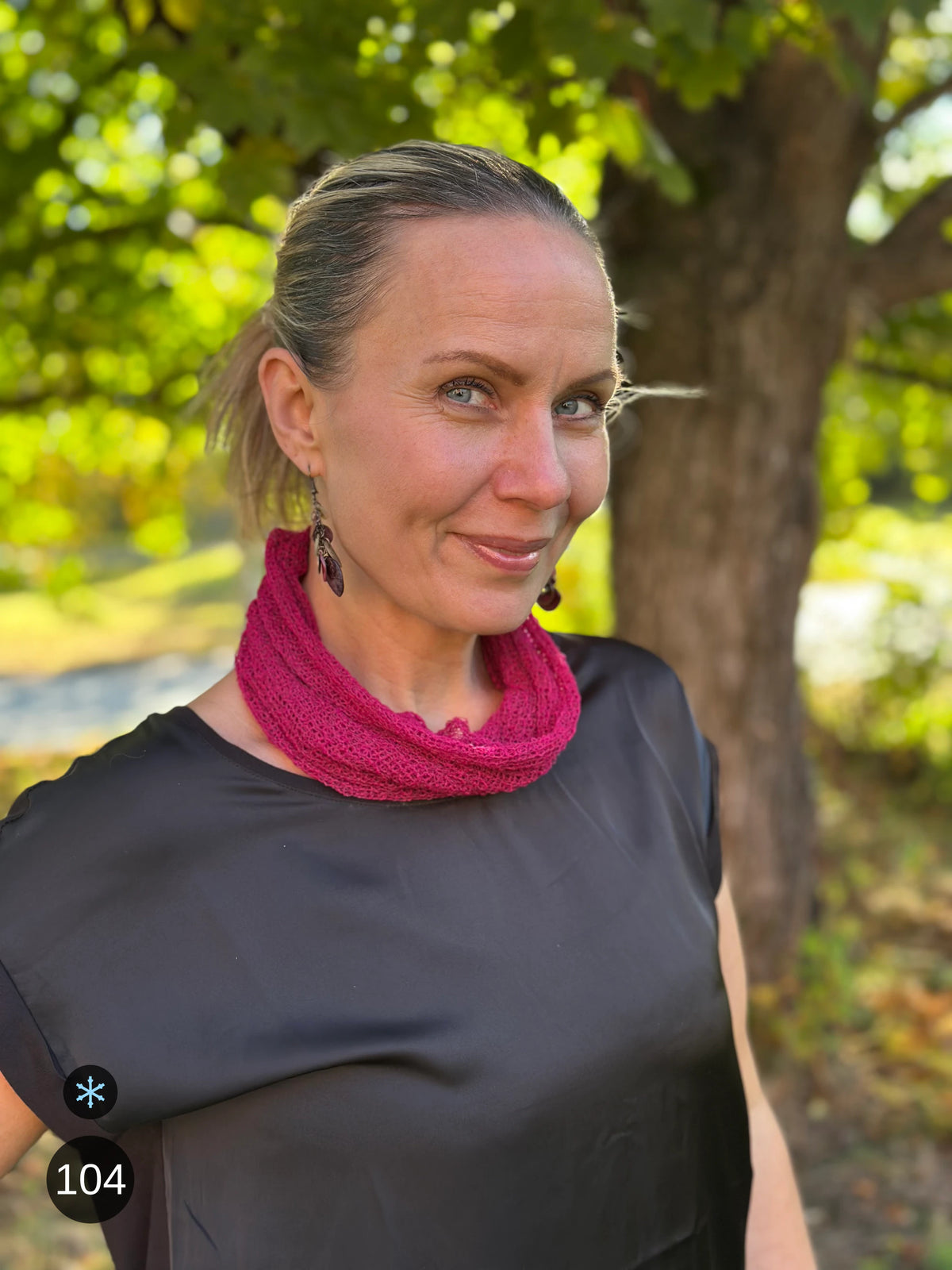 Kvinna bär Eva Tubsjal i färg 104 Fuchsia, en kall färg i krispig blåröd fuchsia, liten finstickad tubsjal buren som krage, fotad i höstsol under en lönn. Ulla Jacobsson Kläder.