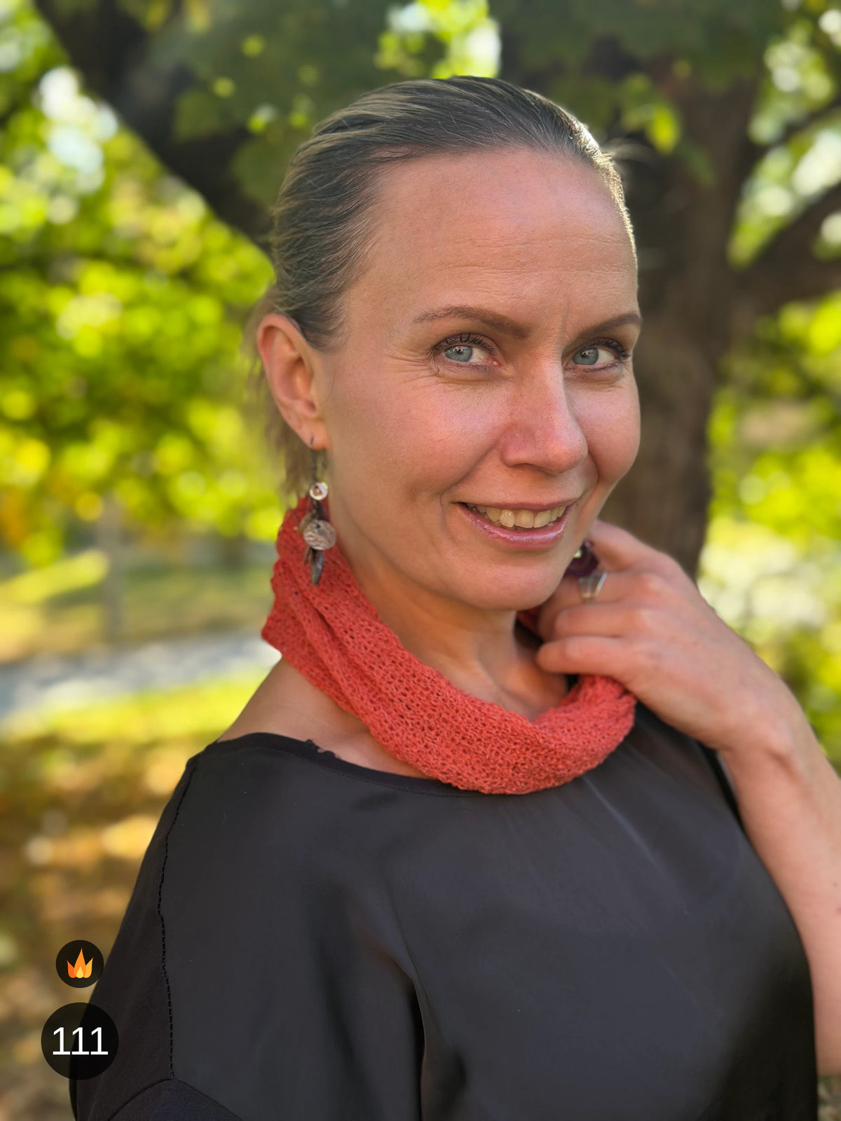 Kvinna bär Eva Tubsjal i färg 111 Rost, en varm färg i roströd tegelton, liten finstickad tubsjal buren som krage, fotad i höstsol under en lönn. Ulla Jacobsson Kläder.