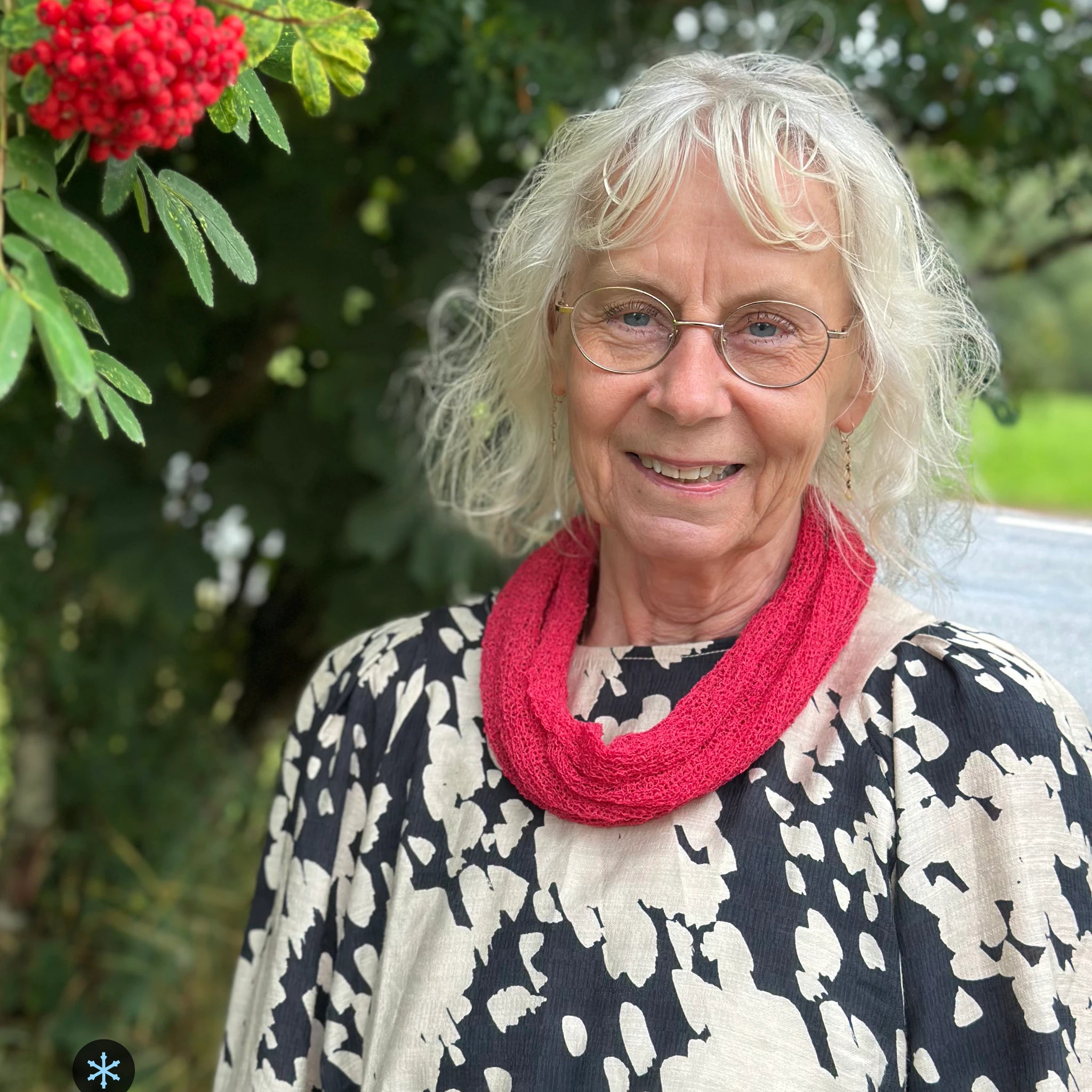 Kvinna bär Eva Tubsjal i färg 116 Azaleröd, en kall färg i klar azaleröd med hallonton, liten finstickad tubsjal buren mjukt runt halsen, fotad utomhus i grönska. Ulla Jacobsson Kläder.