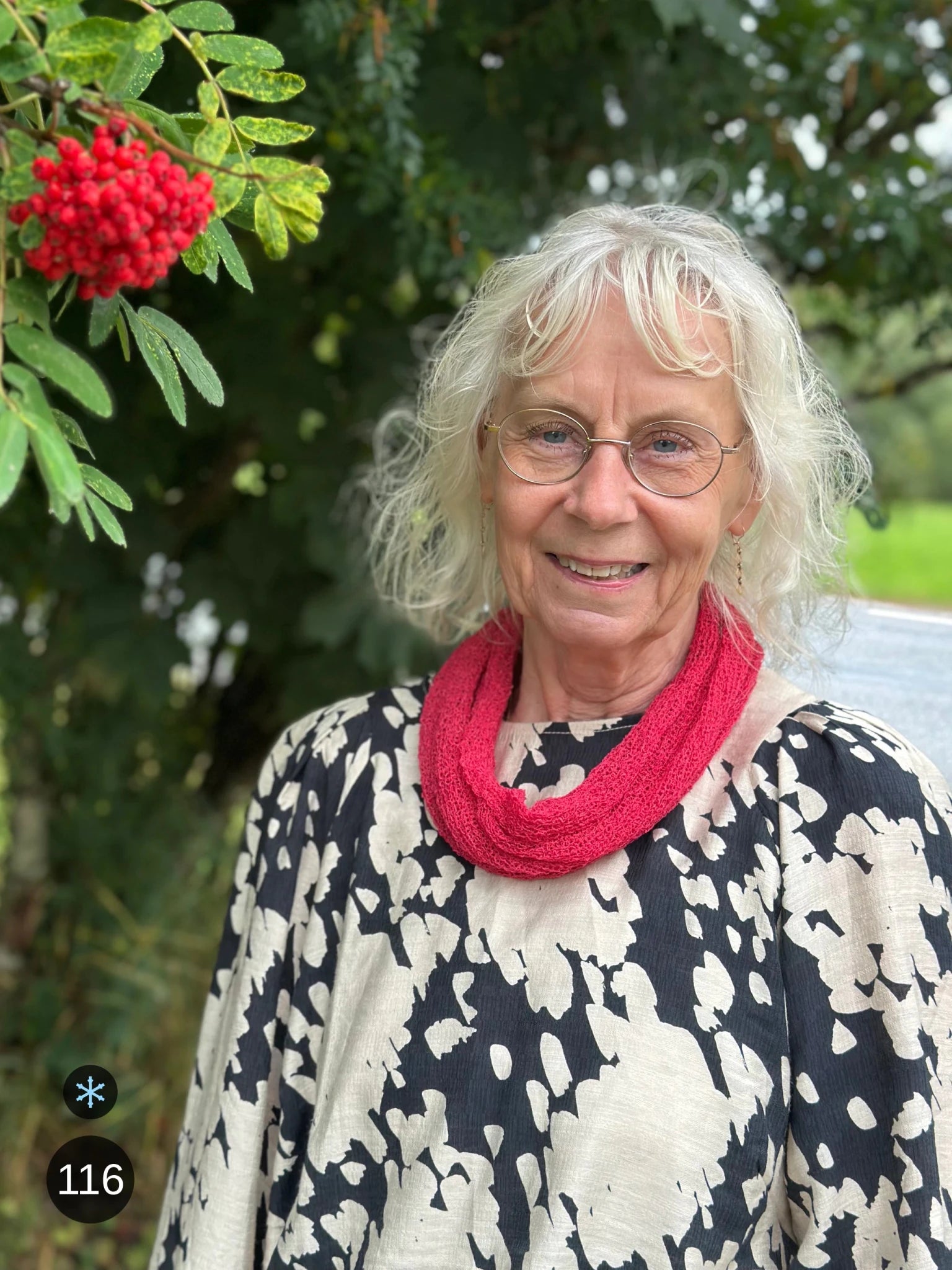 Kvinna bär Eva Tubsjal i färg 116 Azaleröd, en kall färg i klar azaleröd med hallonton, liten finstickad tubsjal buren mjukt runt halsen, fotad utomhus i grönska. Ulla Jacobsson Kläder.