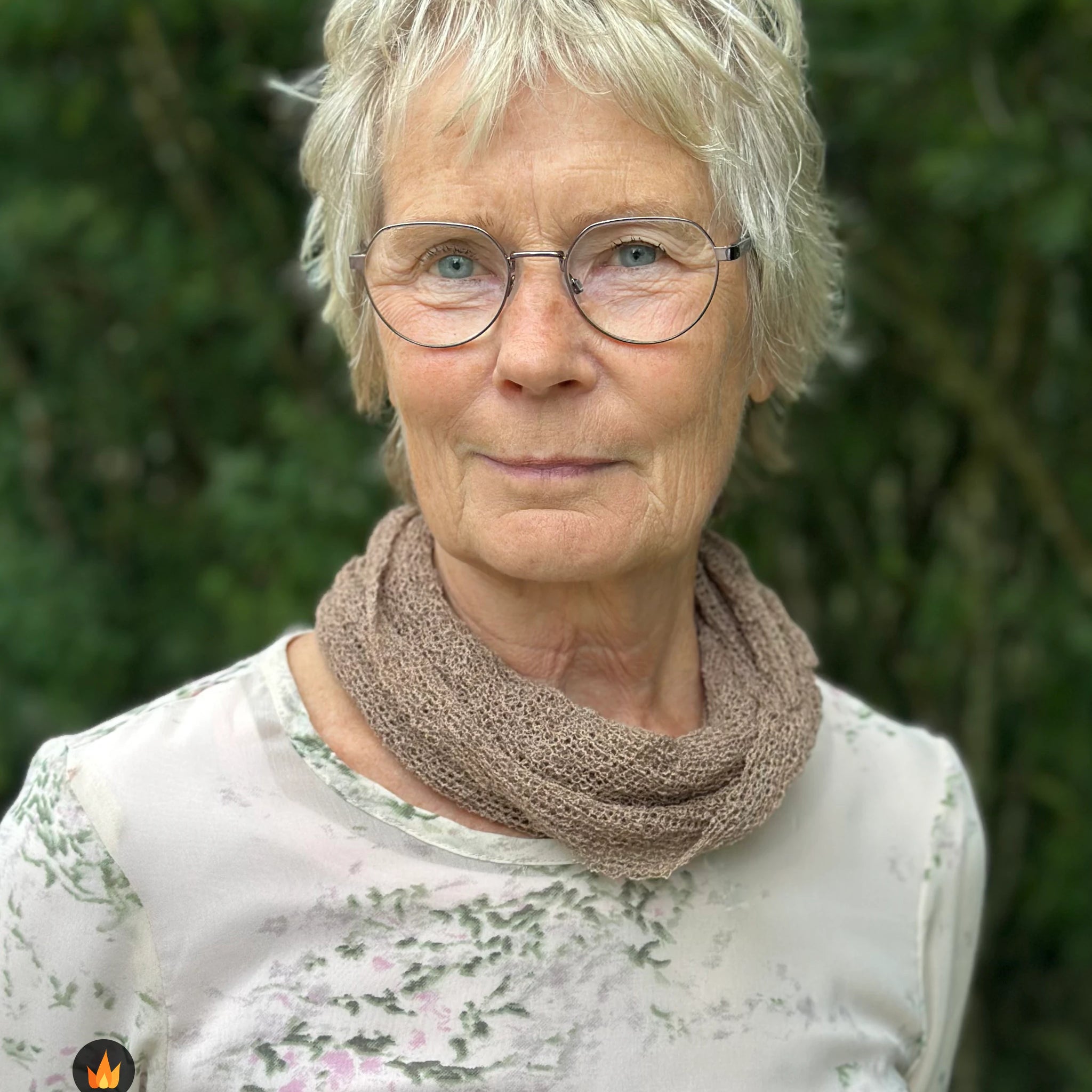 Kvinna bär Eva Tubsjal i färg 119 Cashmere, en varm färg i mjuk gråbeige cashmereton, liten finstickad tubsjal buren mjukt runt halsen, fotad utomhus i grönska. Ulla Jacobsson Kläder.