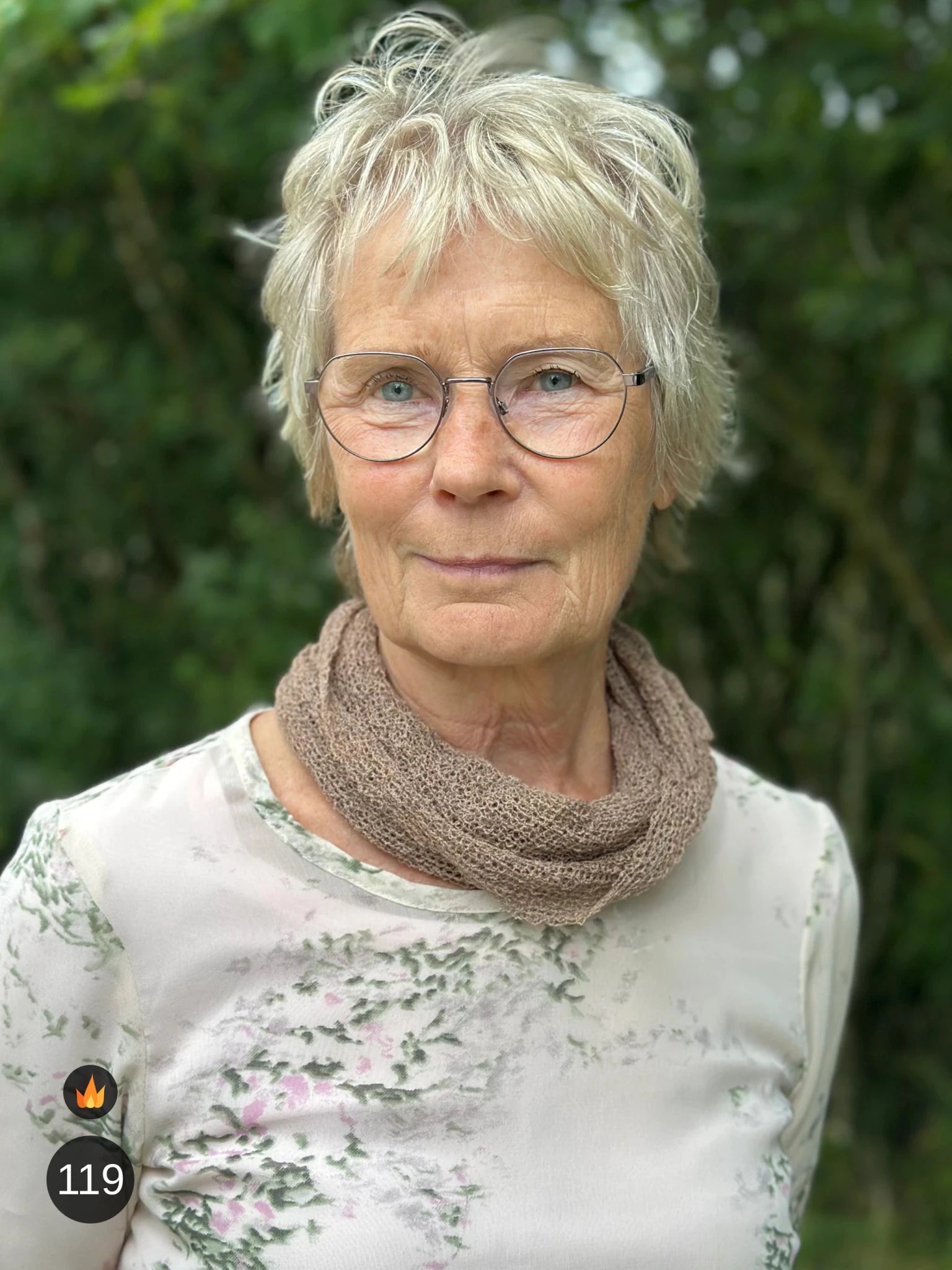 Kvinna bär Eva Tubsjal i färg 119 Cashmere, en varm färg i mjuk gråbeige cashmereton, liten finstickad tubsjal buren mjukt runt halsen, fotad utomhus i grönska. Ulla Jacobsson Kläder.