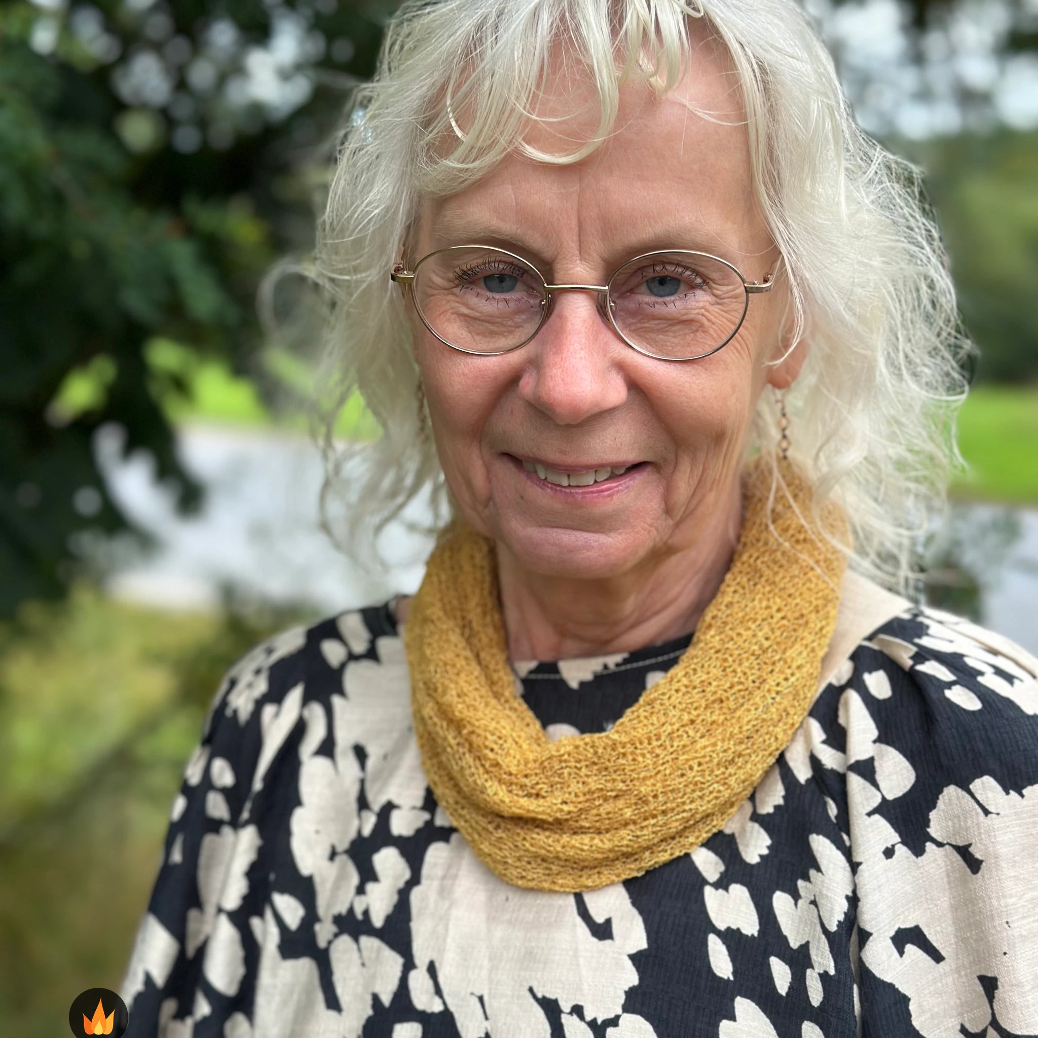 Kvinna bär Eva Tubsjal i färg 121 Ljus senap, en varm färg i ljus senapsgul, liten finstickad tubsjal buren mjukt runt halsen, fotad utomhus i grönska. Ulla Jacobsson Kläder.