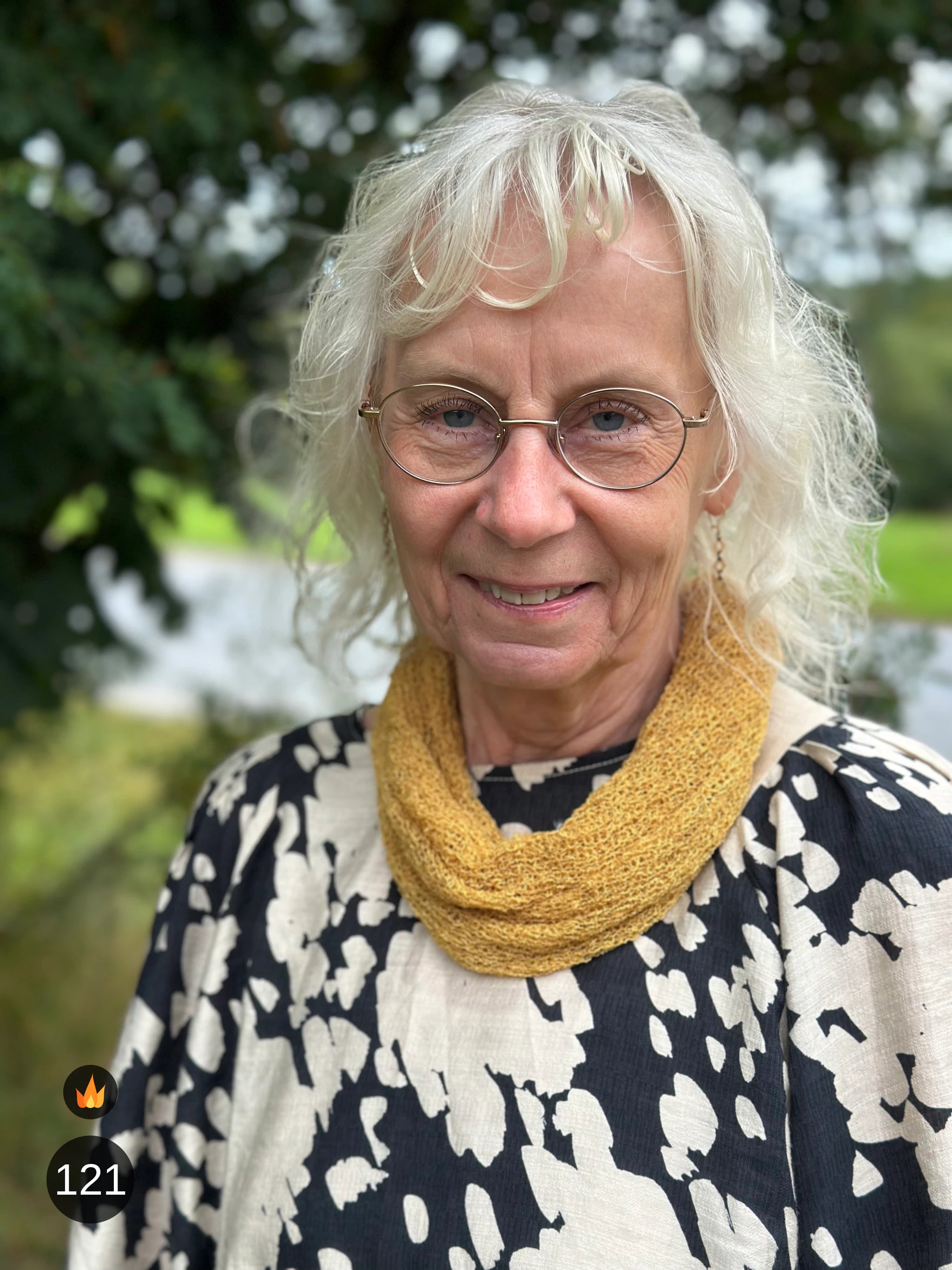 Kvinna bär Eva Tubsjal i färg 121 Ljus senap, en varm färg i ljus senapsgul, liten finstickad tubsjal buren mjukt runt halsen, fotad utomhus i grönska. Ulla Jacobsson Kläder.