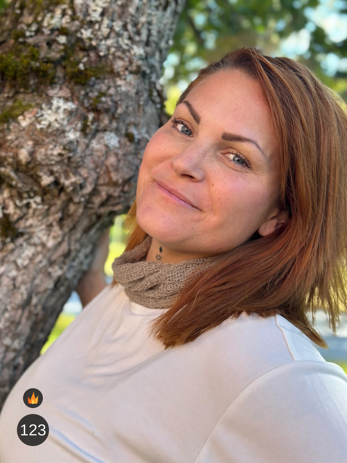 Kvinna bär Eva Tubsjal i färg 123 Ljusbeige, en varm färg i ljusbeige med varm sandton, liten finstickad tubsjal buren som krage, fotad vid en mossig trädstam i höstsol. Ulla Jacobsson Kläder.