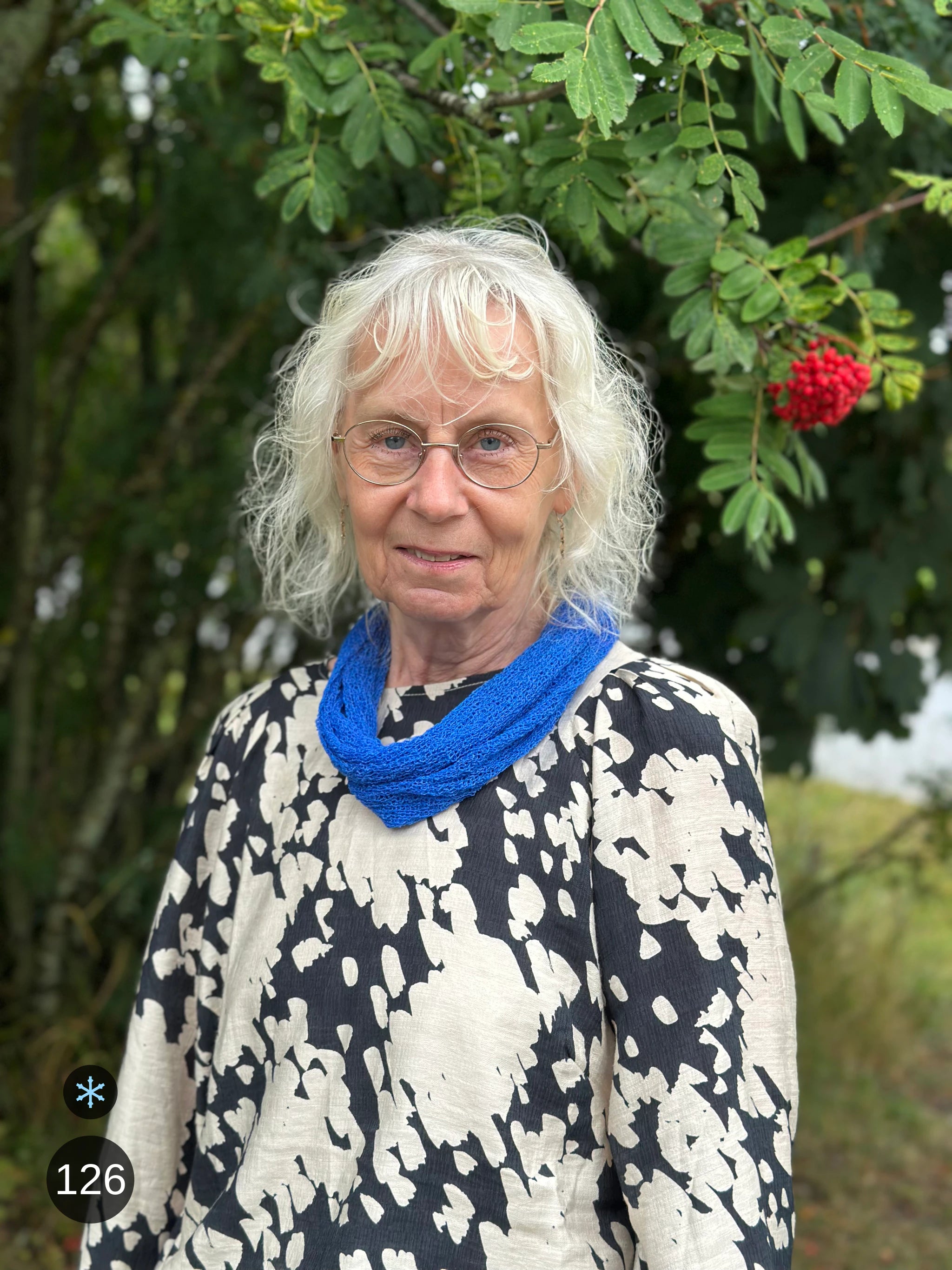 Kvinna bär Eva Tubsjal i färg 126 Sverigeblå, en kall färg i klar sverigeblå, liten finstickad tubsjal buren mjukt runt halsen, fotad under en rönn med röda bär. Ulla Jacobsson Kläder.