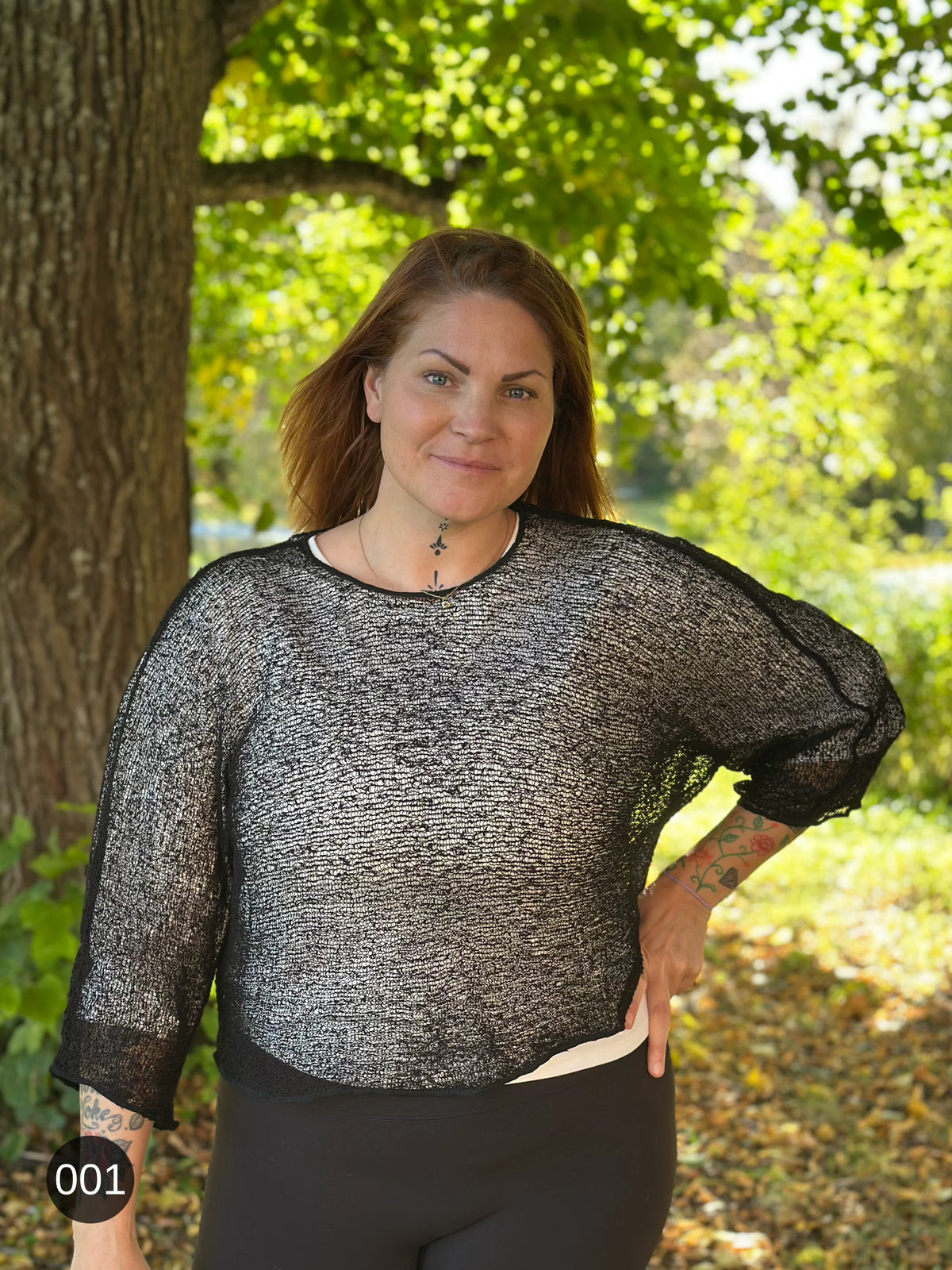 Kvinna bär Lebar Poncho i färg 001 Svart, en neutral djupsvart med matt yta, finstickad poncho med vida ärmar som faller luftigt, fotad under en lönn i höstsol. Ulla Jacobsson Kläder.