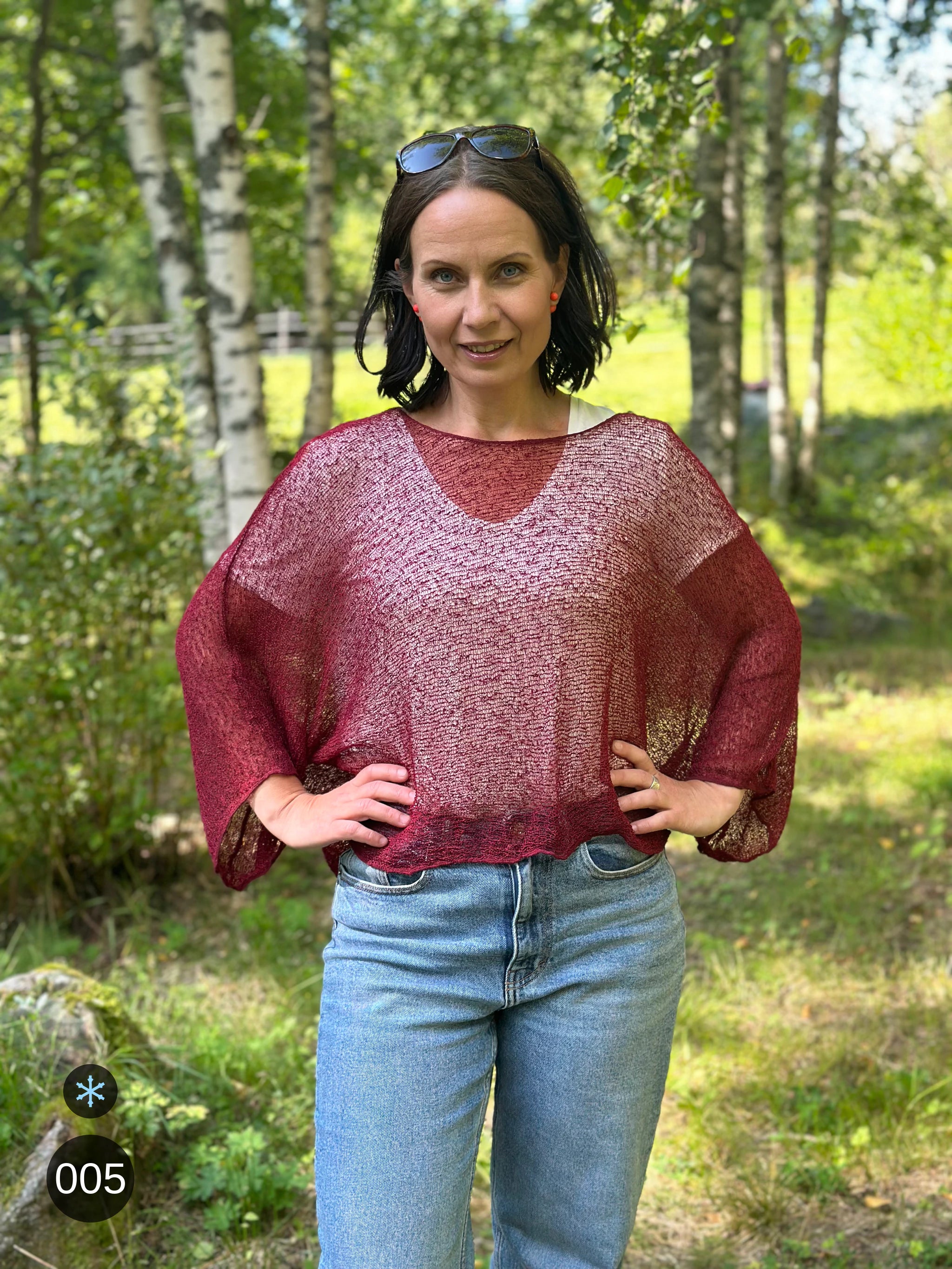 Kvinna bär Lebar Poncho i färg 005 Vinröd, en kall färg i dämpad vinröd, finstickad poncho med vida ärmar som faller luftigt, fotad i björkskog i sommargrönska. Ulla Jacobsson Kläder.