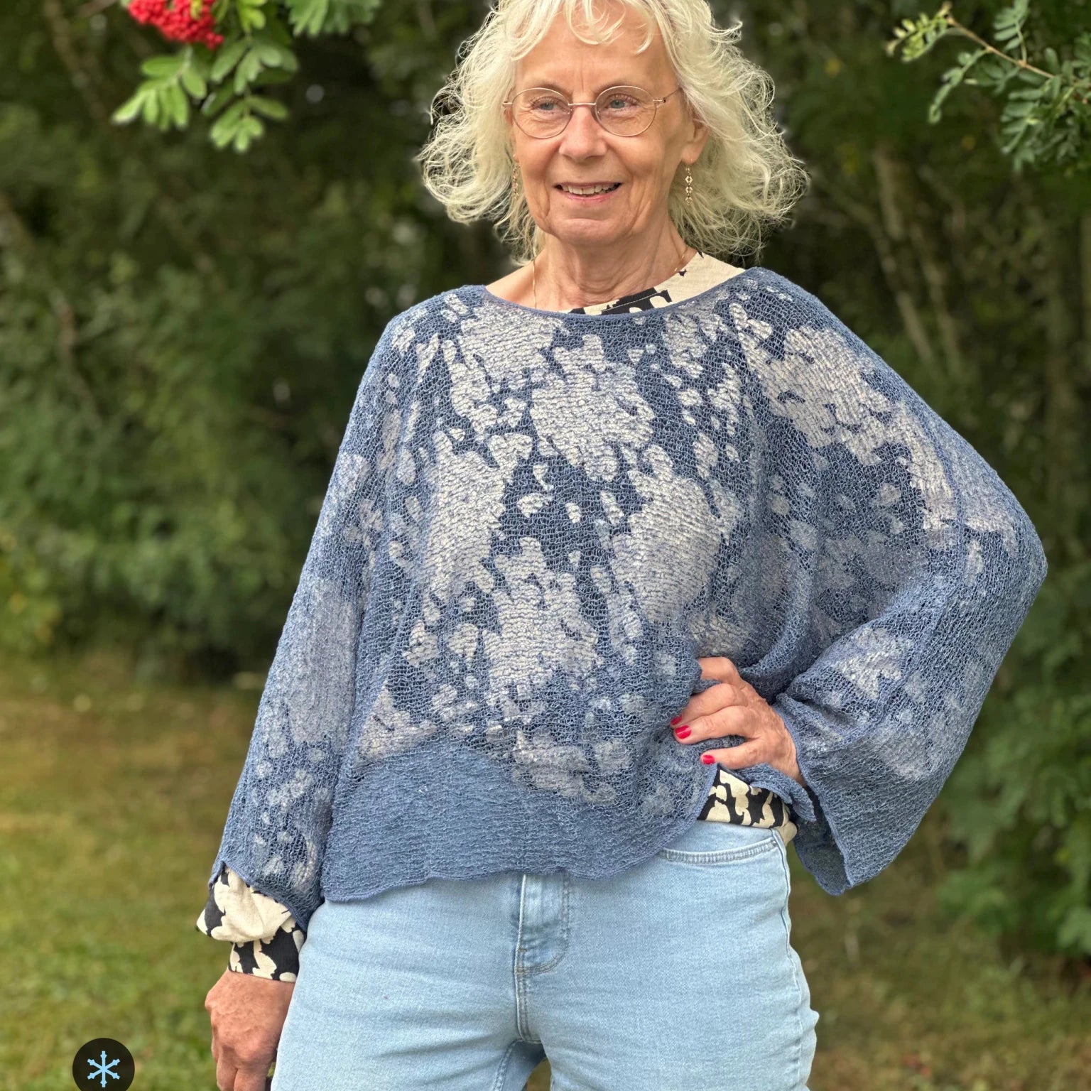 Kvinna bär Lebar Poncho i färg 025 Jeansblå, en kall färg i dämpad denimblå, finstickad poncho med vida ärmar som faller luftigt, fotad utomhus i grönska. Ulla Jacobsson Kläder.