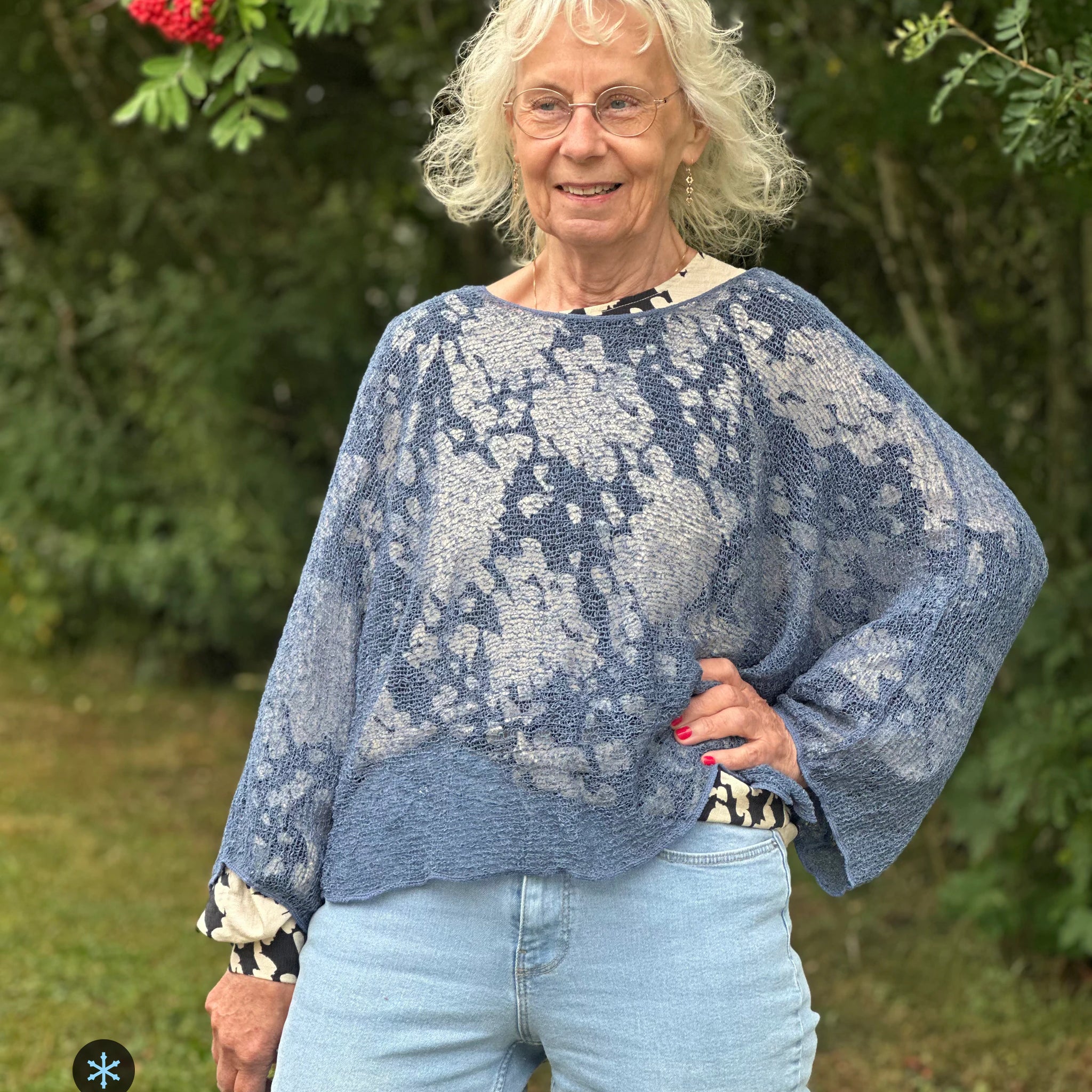 Kvinna bär Lebar Poncho i färg 025 Jeansblå, en kall färg i dämpad denimblå, finstickad poncho med vida ärmar som faller luftigt, fotad utomhus i grönska. Ulla Jacobsson Kläder.