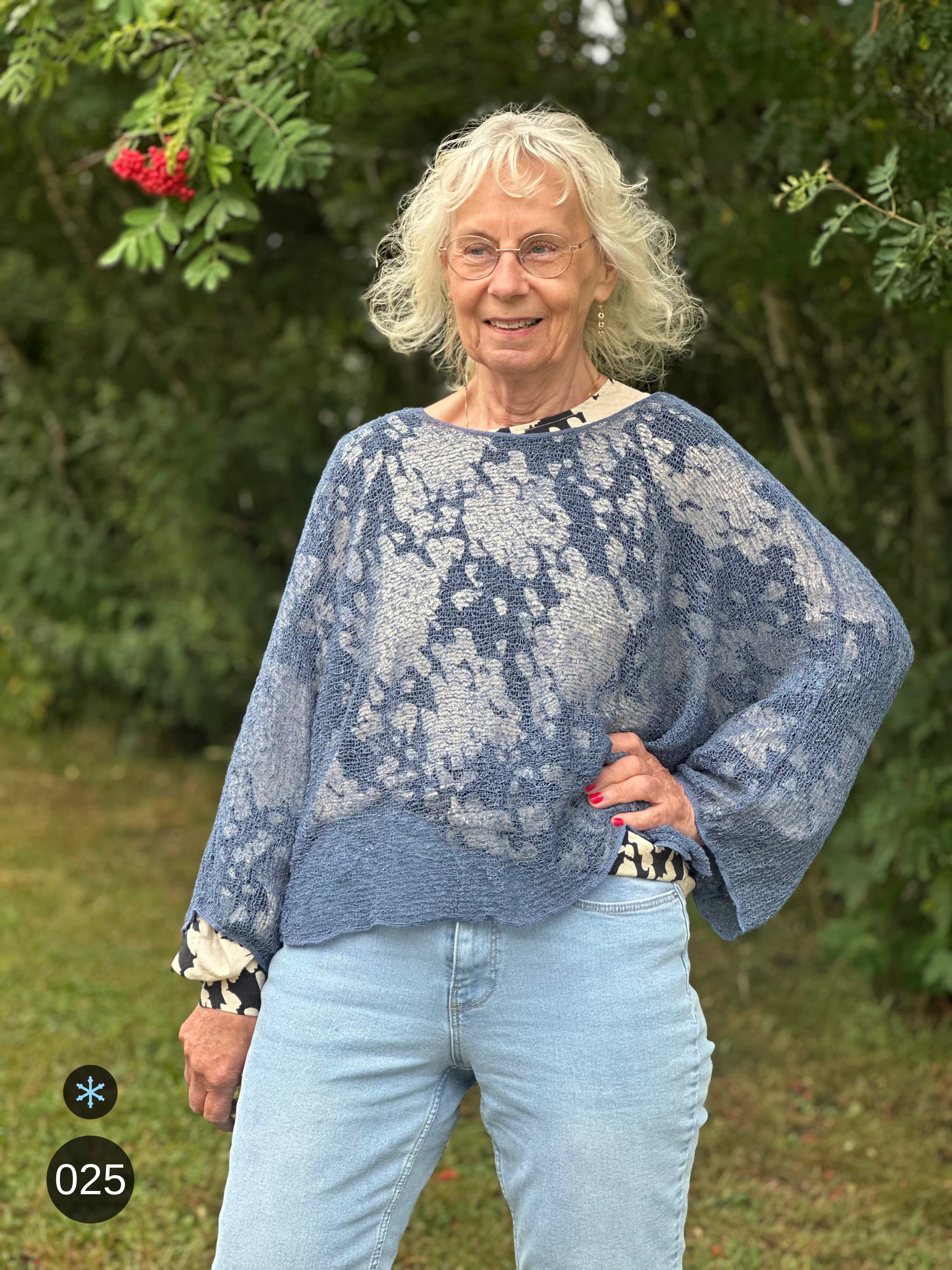 Kvinna bär Lebar Poncho i färg 025 Jeansblå, en kall färg i dämpad denimblå, finstickad poncho med vida ärmar som faller luftigt, fotad utomhus i grönska. Ulla Jacobsson Kläder.