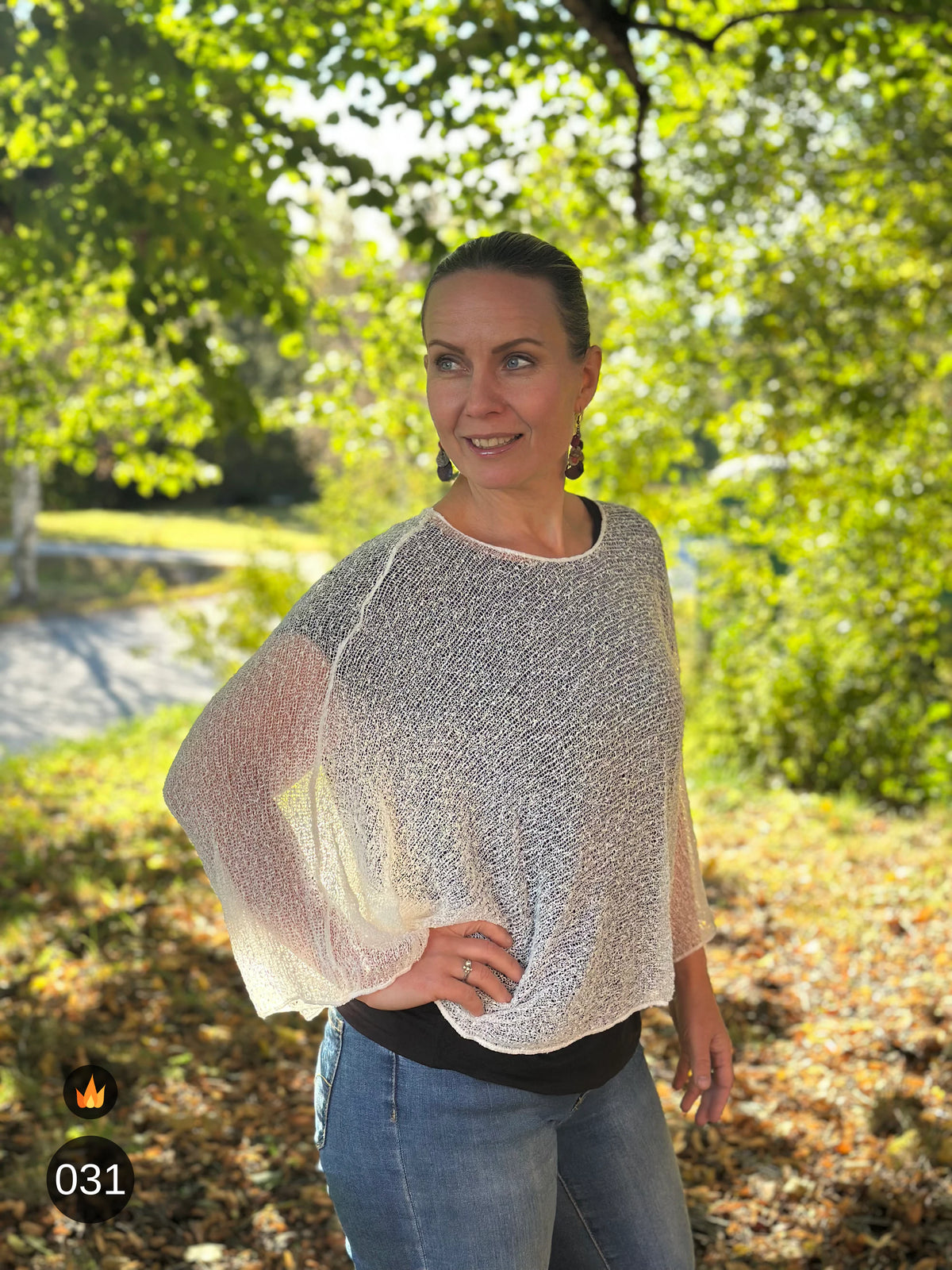 Kvinna bär Lebar Poncho i färg 031 Offwhite, en varm färg i mjuk naturvit med gräddig underton, finstickad poncho med vida ärmar som faller luftigt, fotad i en park i höstsol. Ulla Jacobsson Kläder.