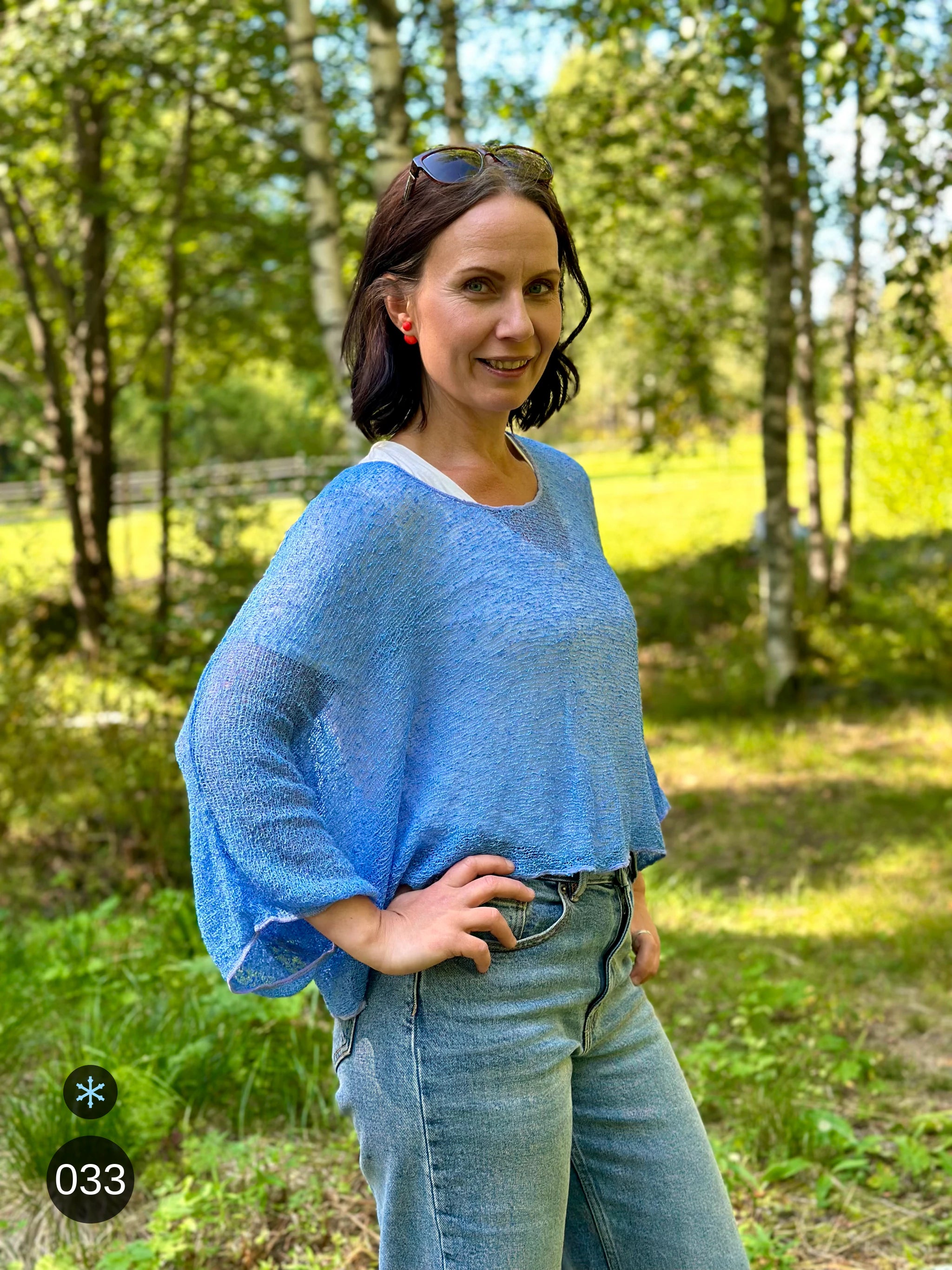 Kvinna bär Lebar Poncho i färg 033 Ljusblå, en kall färg i ljus himmelsblå, finstickad poncho med vida ärmar som faller luftigt, fotad i björkskog i sommarljus. Ulla Jacobsson Kläder.