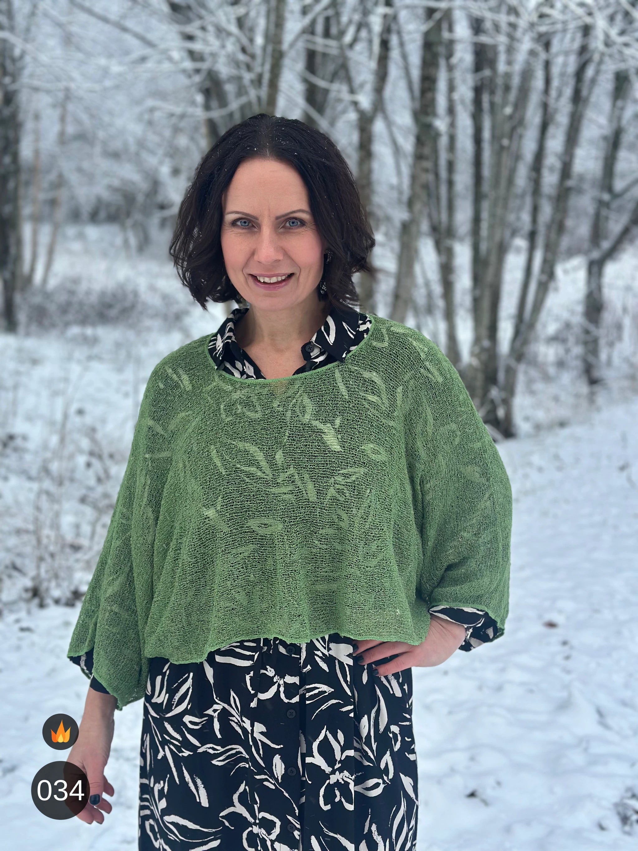 Kvinna bär Lebar Poncho i färg 034 Ljusgrön, en varm färg i frisk ljusgrön, finstickad poncho med vida ärmar som faller luftigt, fotad i snöig björkskog. Ulla Jacobsson Kläder.