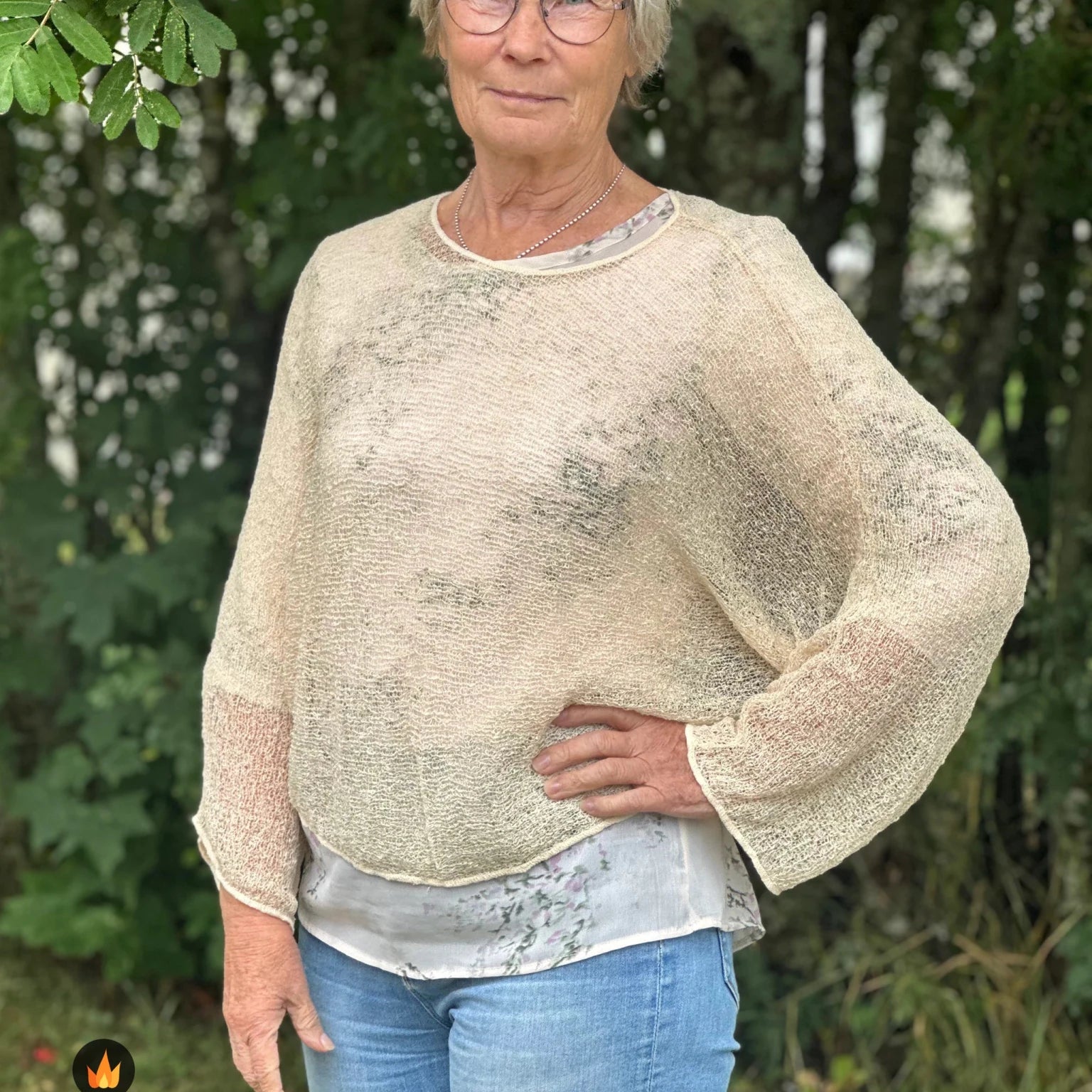 Kvinna bär Lebar Poncho i färg 036 Ljus sand, en varm färg i ljus sandbeige, finstickad poncho med vida ärmar som faller luftigt, fotad utomhus i grönska. Ulla Jacobsson Kläder.