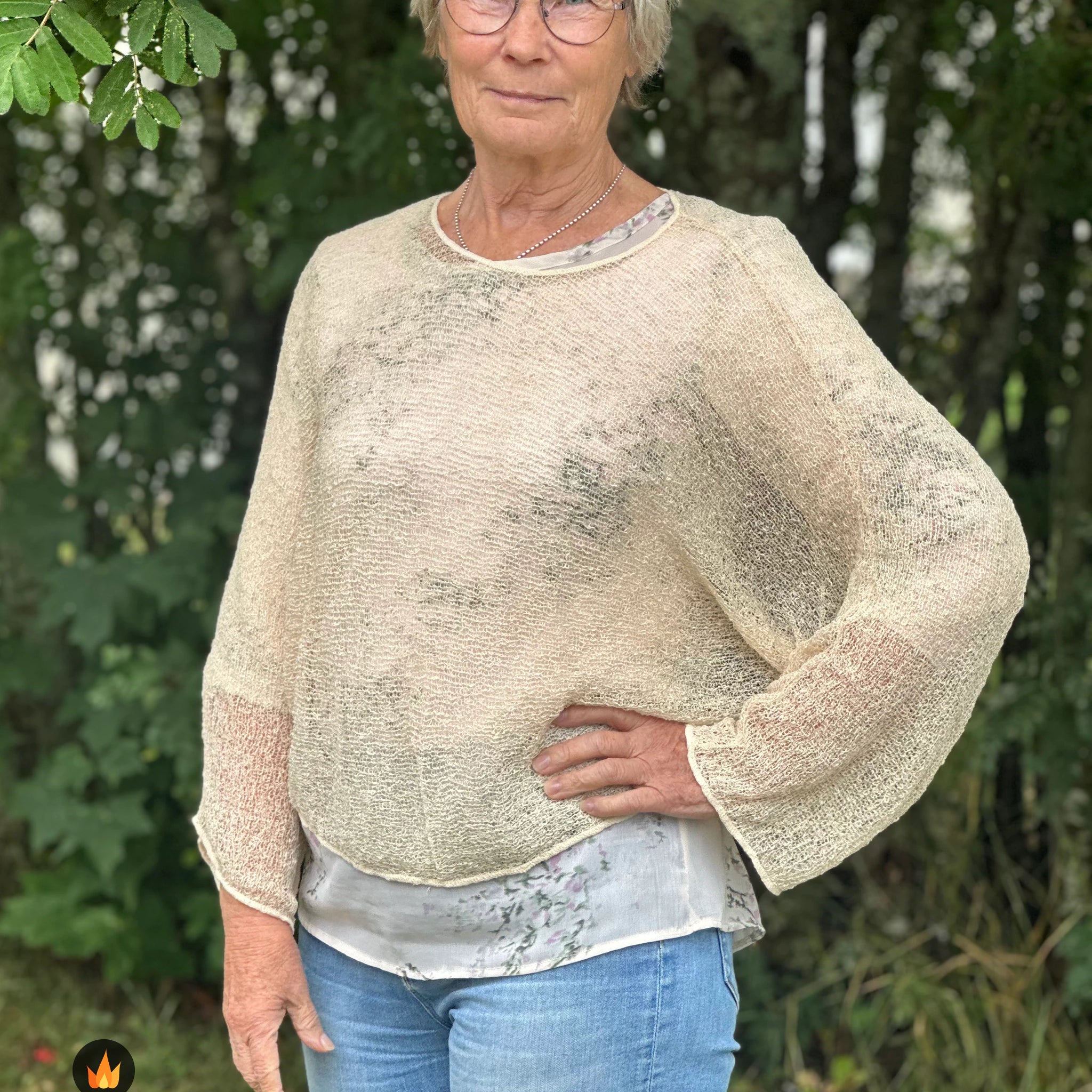 Kvinna bär Lebar Poncho i färg 036 Ljus sand, en varm färg i ljus sandbeige, finstickad poncho med vida ärmar som faller luftigt, fotad utomhus i grönska. Ulla Jacobsson Kläder.