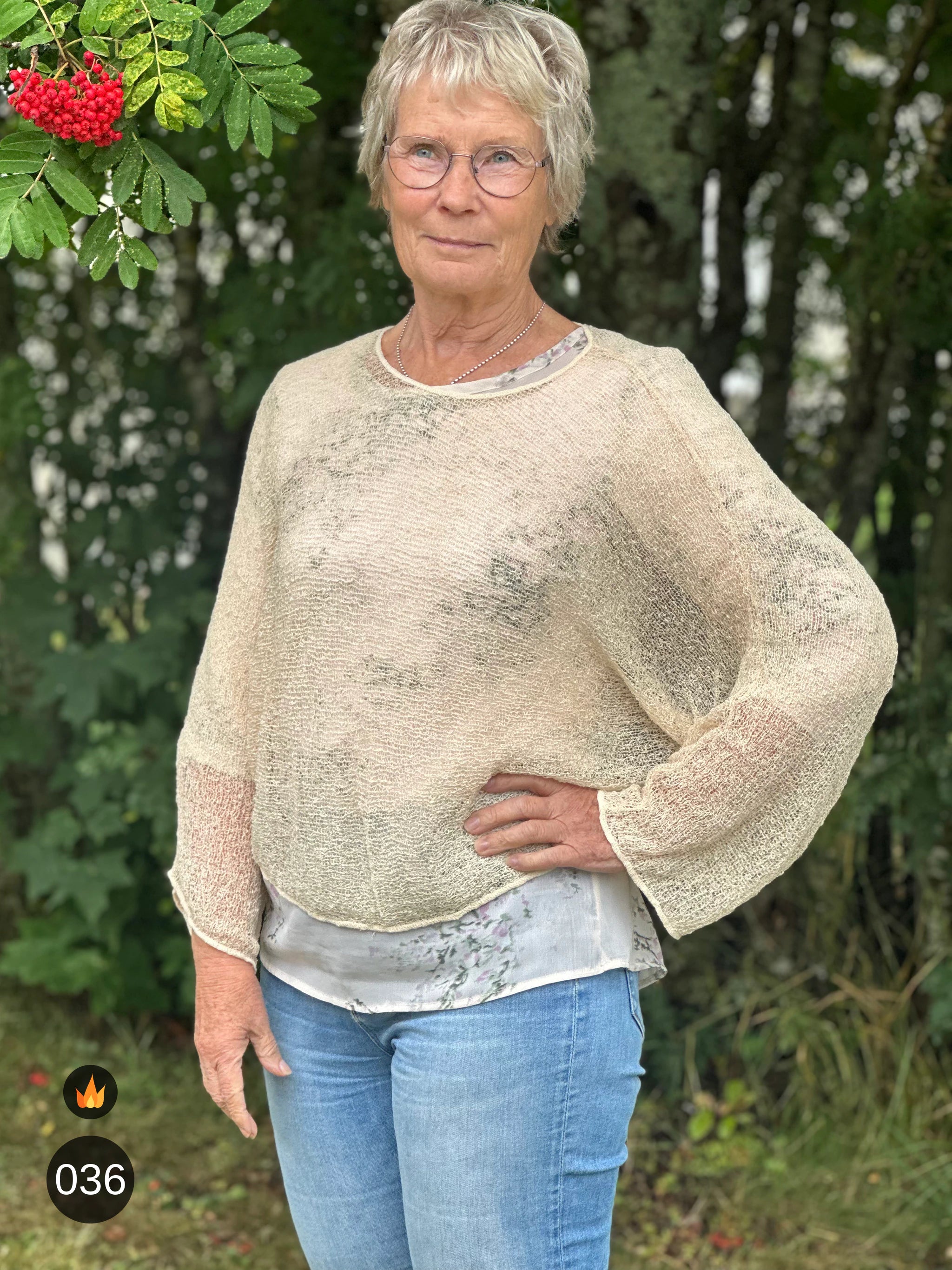 Kvinna bär Lebar Poncho i färg 036 Ljus sand, en varm färg i ljus sandbeige, finstickad poncho med vida ärmar som faller luftigt, fotad utomhus i grönska. Ulla Jacobsson Kläder.