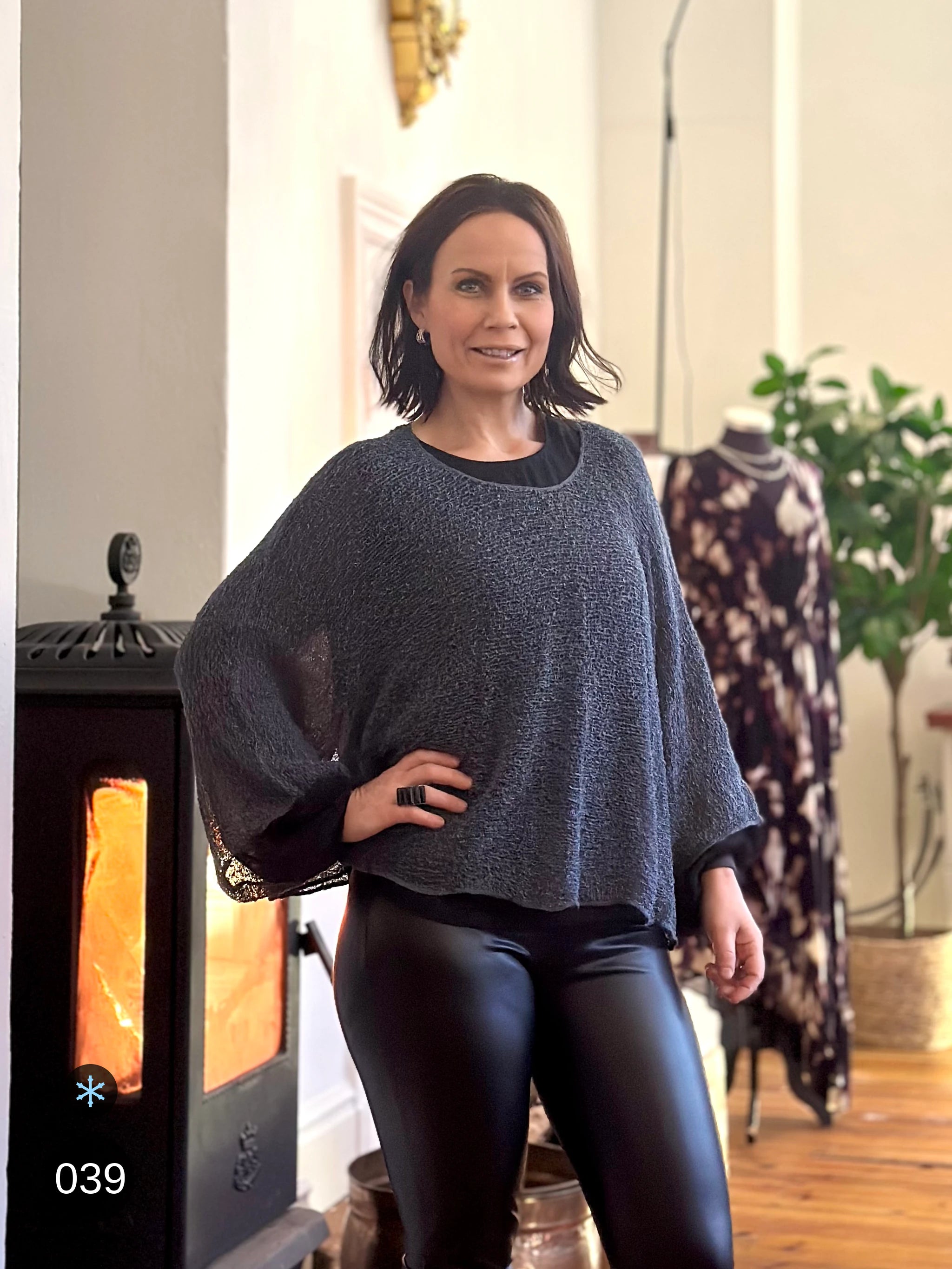 Kvinna bär Lebar Poncho i färg 039 Anilin, en kall färg i gråblå melerad ton, finstickad poncho med vida ärmar som faller luftigt, fotad inomhus i butiken framför en brasa. Ulla Jacobsson Kläder.