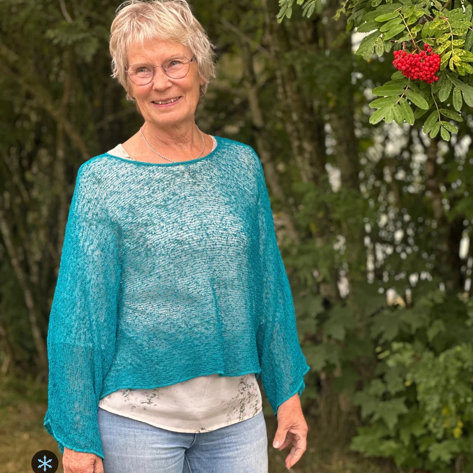 Kvinna bär Lebar Poncho i färg 040 Petrol, en kall färg i djup petrolblå, finstickad poncho med vida ärmar som faller luftigt, fotad utomhus i grönska. Ulla Jacobsson Kläder.