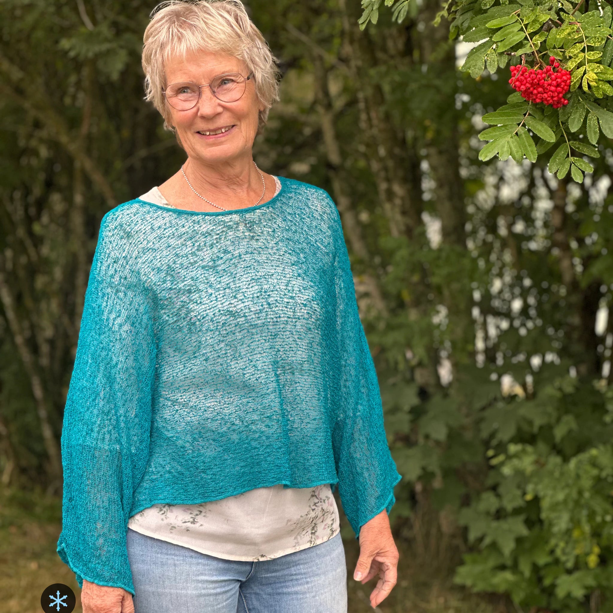 Kvinna bär Lebar Poncho i färg 040 Petrol, en kall färg i djup petrolblå, finstickad poncho med vida ärmar som faller luftigt, fotad utomhus i grönska. Ulla Jacobsson Kläder.