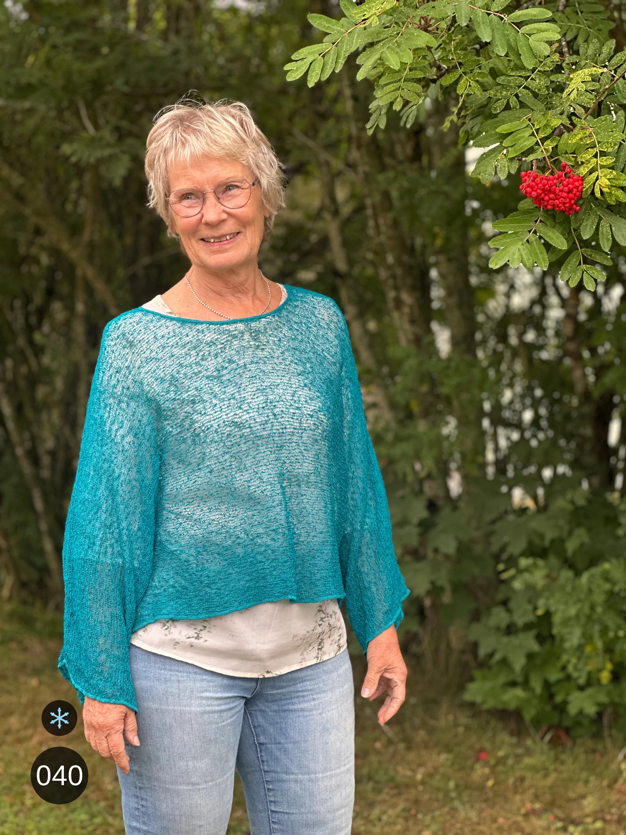 Kvinna bär Lebar Poncho i färg 040 Petrol, en kall färg i djup petrolblå, finstickad poncho med vida ärmar som faller luftigt, fotad utomhus i grönska. Ulla Jacobsson Kläder.