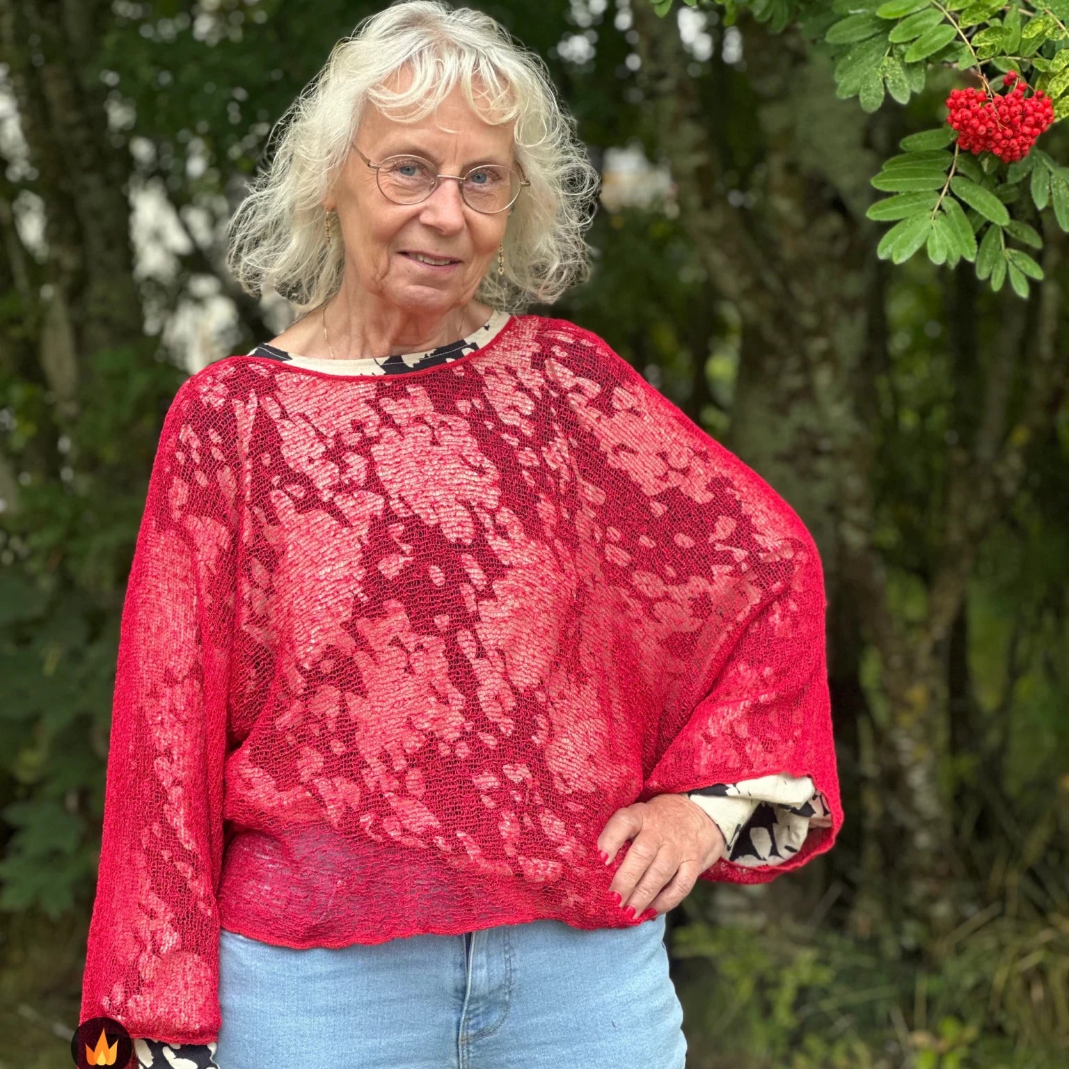 Kvinna bär Lebar Poncho i färg 042 Röd, en varm färg i klar röd, finstickad poncho med vida ärmar som faller luftigt, fotad utomhus i grönska. Ulla Jacobsson Kläder.