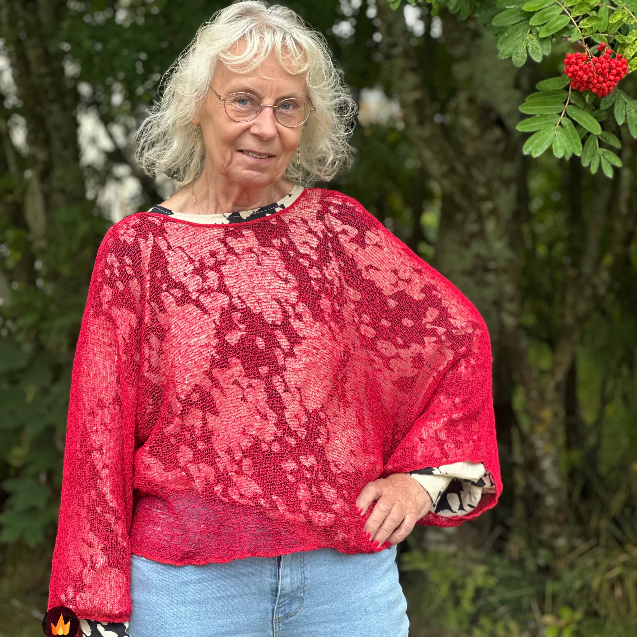 Kvinna bär Lebar Poncho i färg 042 Röd, en varm färg i klar röd, finstickad poncho med vida ärmar som faller luftigt, fotad utomhus i grönska. Ulla Jacobsson Kläder.
