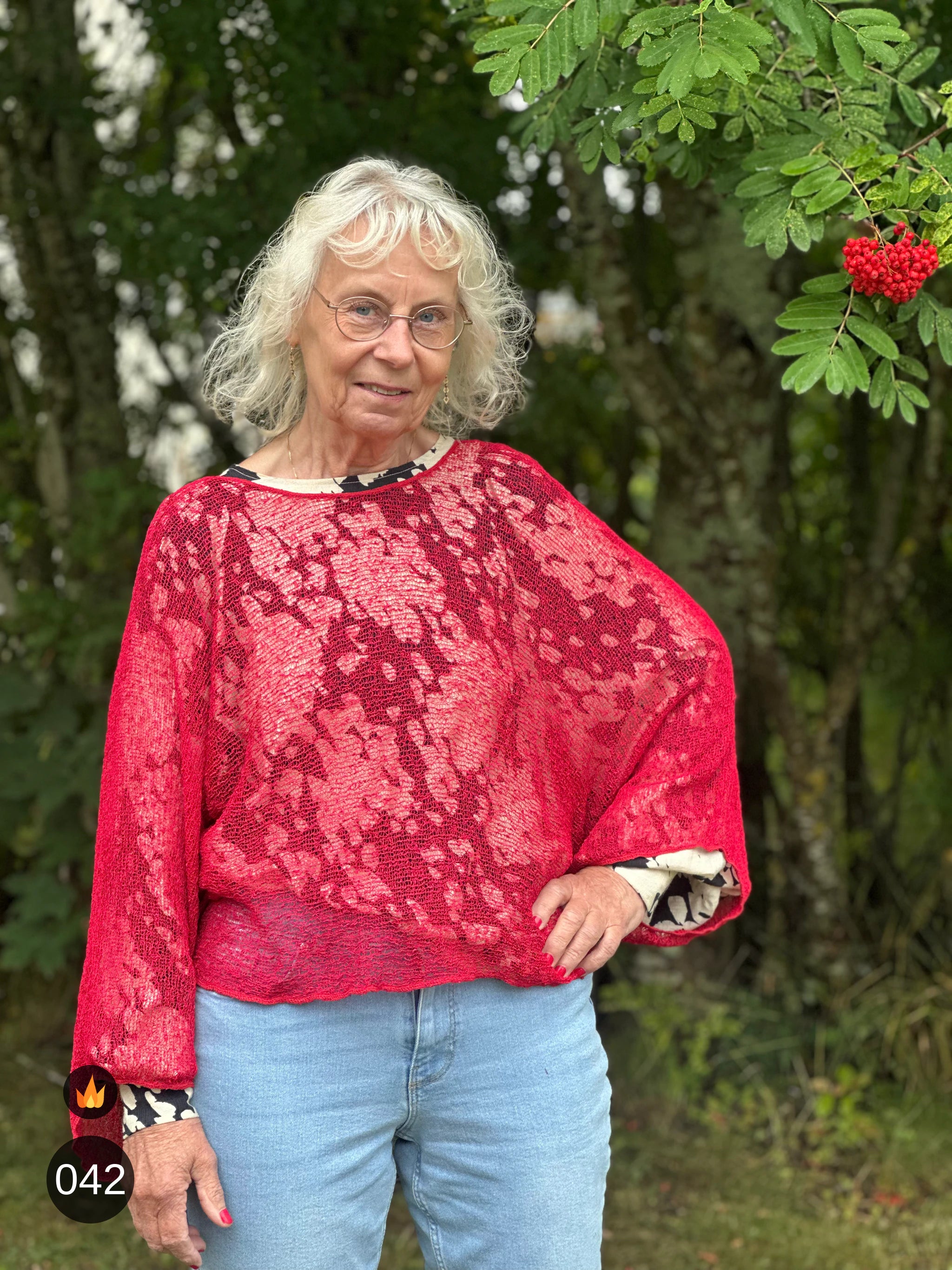 Kvinna bär Lebar Poncho i färg 042 Röd, en varm färg i klar röd, finstickad poncho med vida ärmar som faller luftigt, fotad utomhus i grönska. Ulla Jacobsson Kläder.