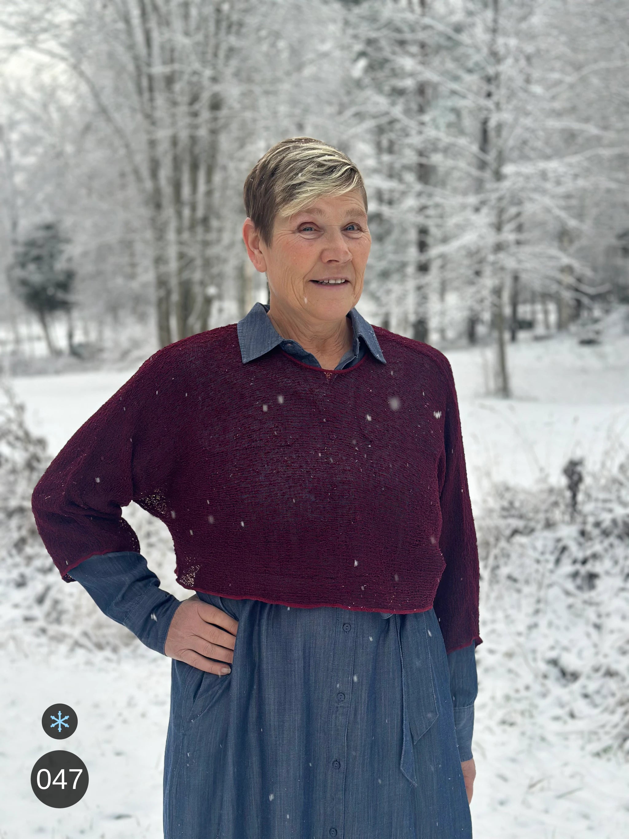 Kvinna bär Lebar Poncho i färg 047 Bourdeaux, en kall färg i djup bourdeauxröd, finstickad poncho med vida ärmar som faller luftigt, fotad i snöig björkskog medan snön faller. Ulla Jacobsson Kläder.