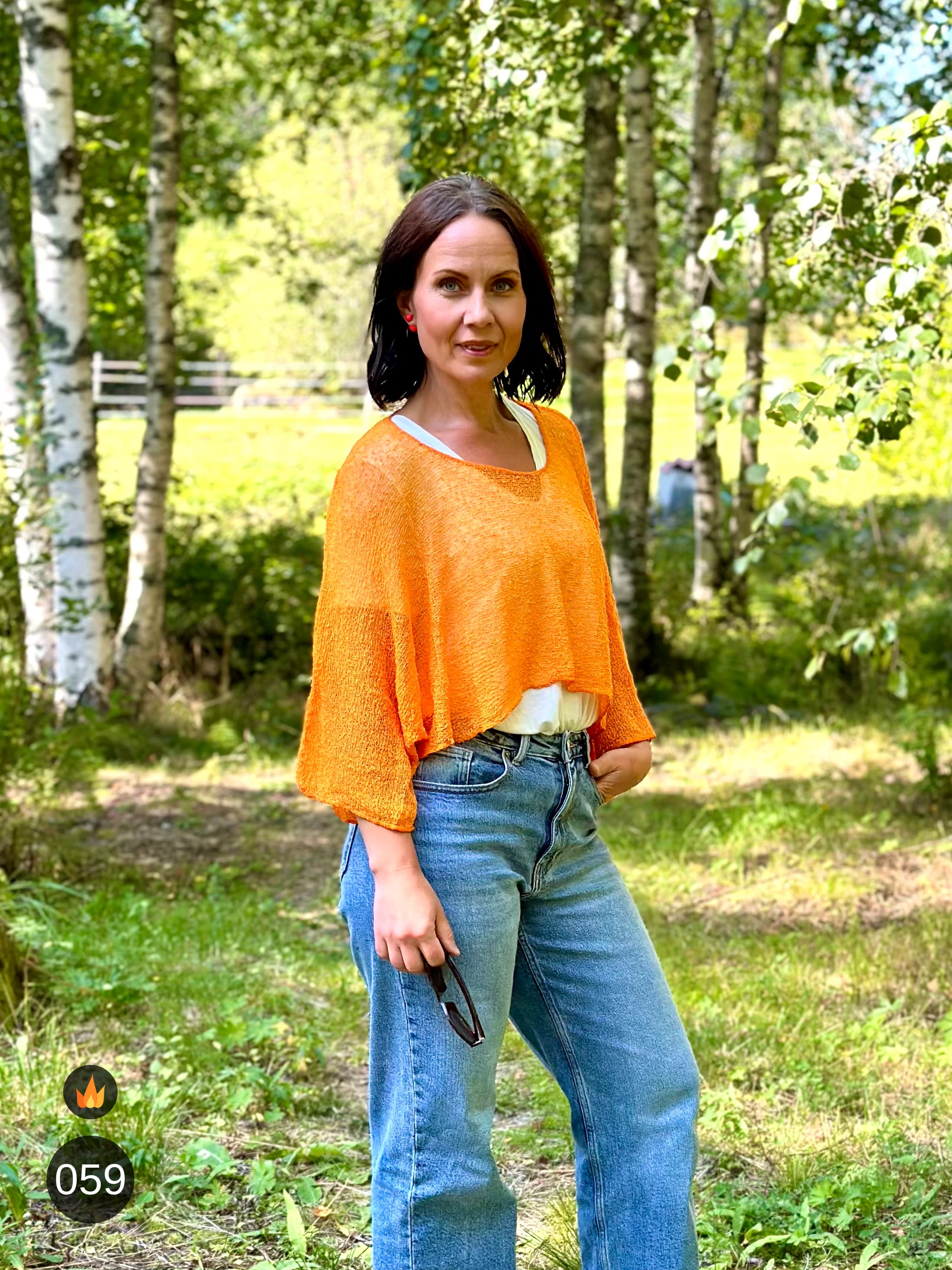 Kvinna bär Lebar Poncho i färg 059 Ljusorange, en varm färg i klar ljusorange, finstickad poncho med vida ärmar som faller luftigt, fotad på gräset i björkskog en sommardag. Ulla Jacobsson Kläder.