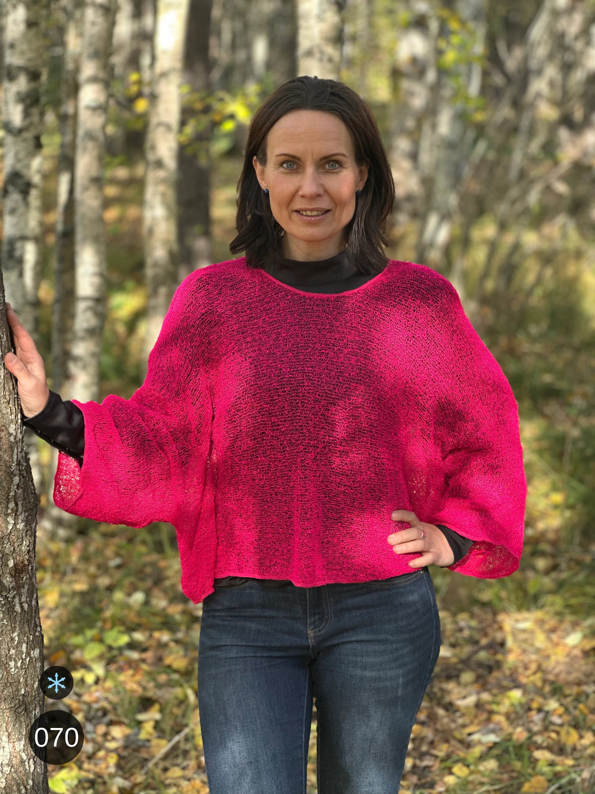 Kvinna bär Lebar Poncho i färg 070 Neonrosa, en kall färg i lysande neonrosa, finstickad poncho med vida ärmar som faller luftigt, fotad i björkskog i höstljus. Ulla Jacobsson Kläder.