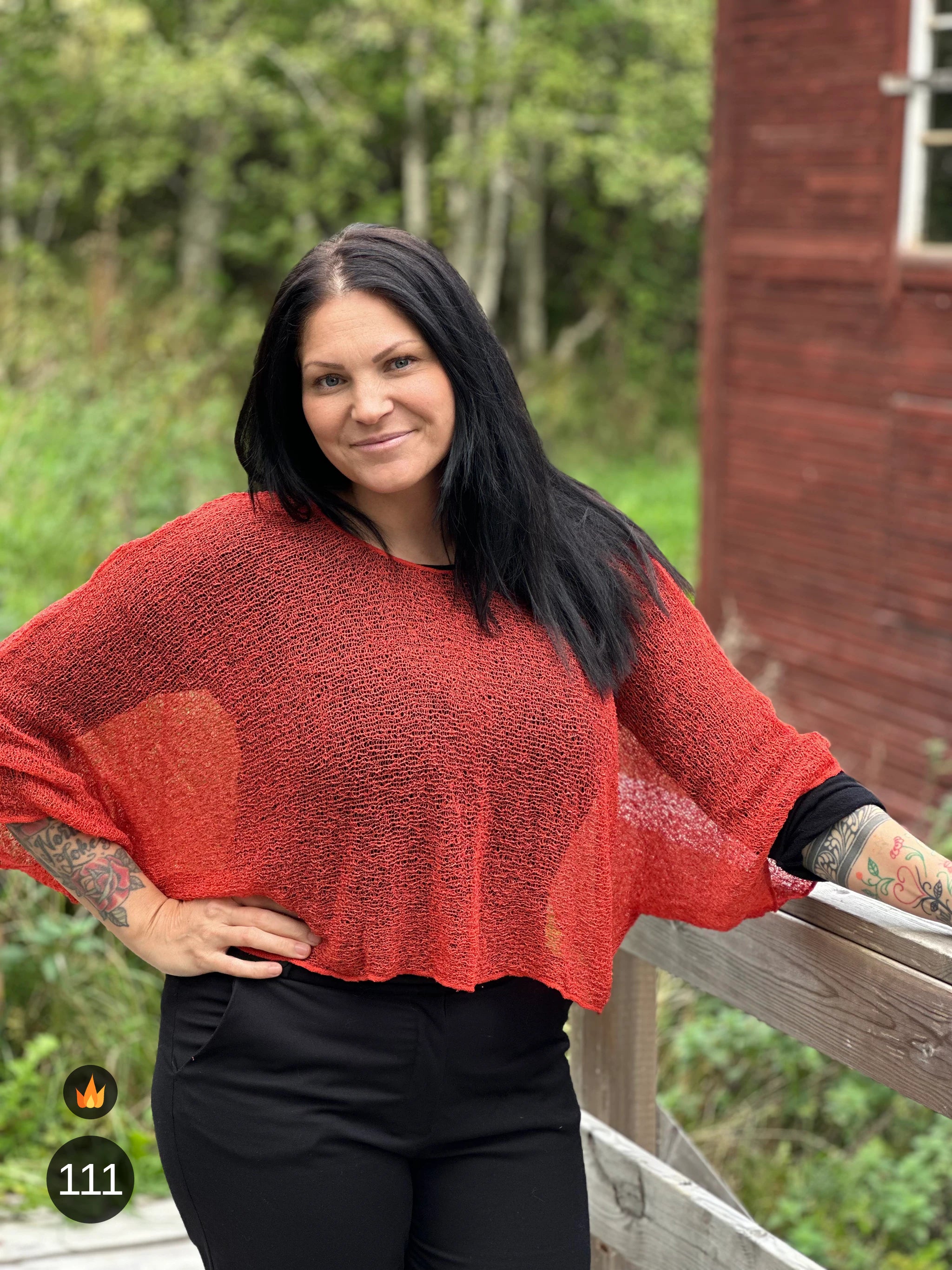 Kvinna bär Lebar Poncho i färg 111 Rost, en varm färg i roströd tegelton, finstickad poncho med vida ärmar som faller luftigt, fotad vid ett trästaket framför ett rött hus. Ulla Jacobsson Kläder.