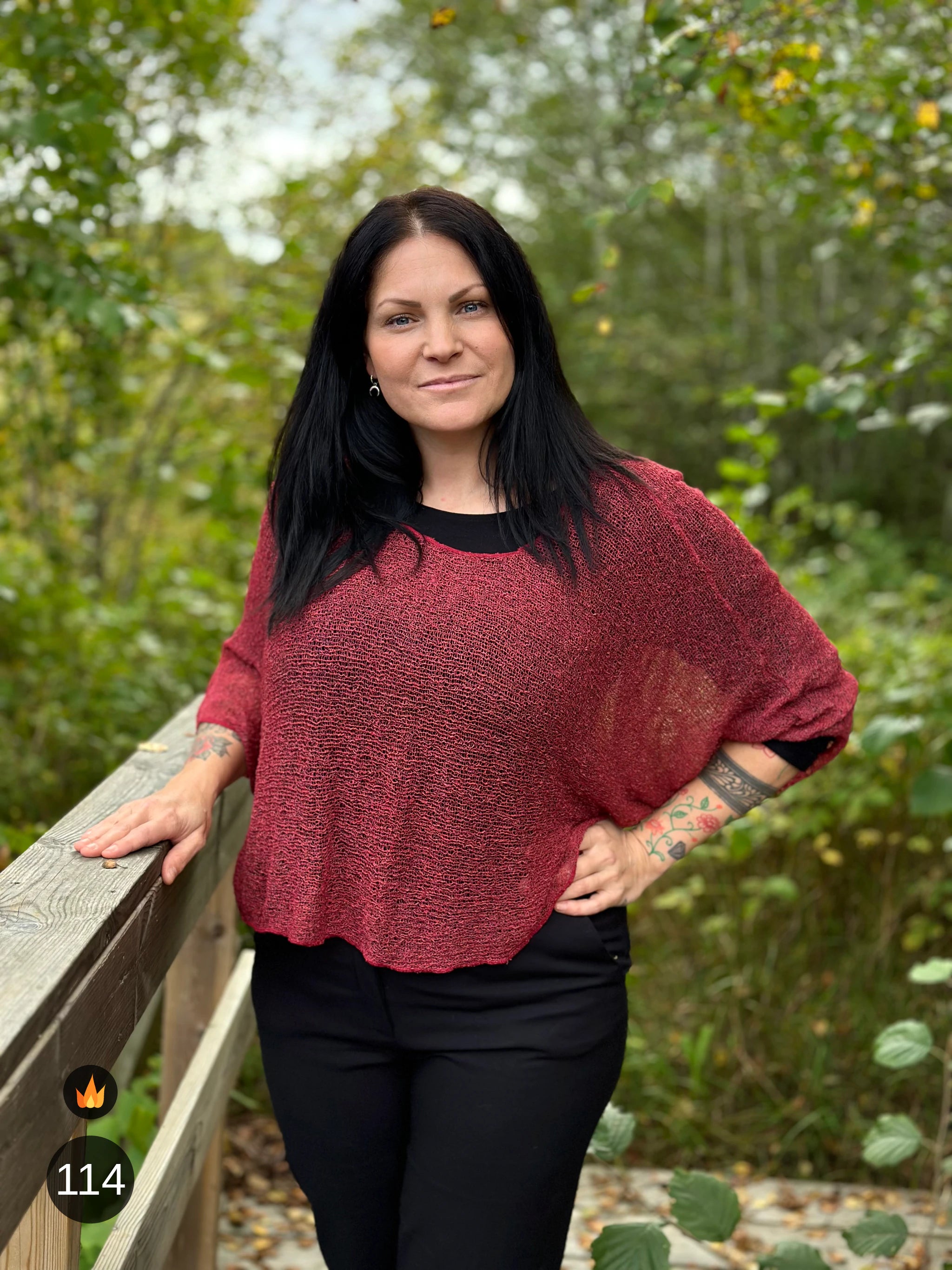 Kvinna bär Lebar Poncho i färg 114 Maroon, en varm färg i dämpad maroon, finstickad poncho med vida ärmar som faller luftigt, fotad vid ett trästaket med skogen bakom. Ulla Jacobsson Kläder.