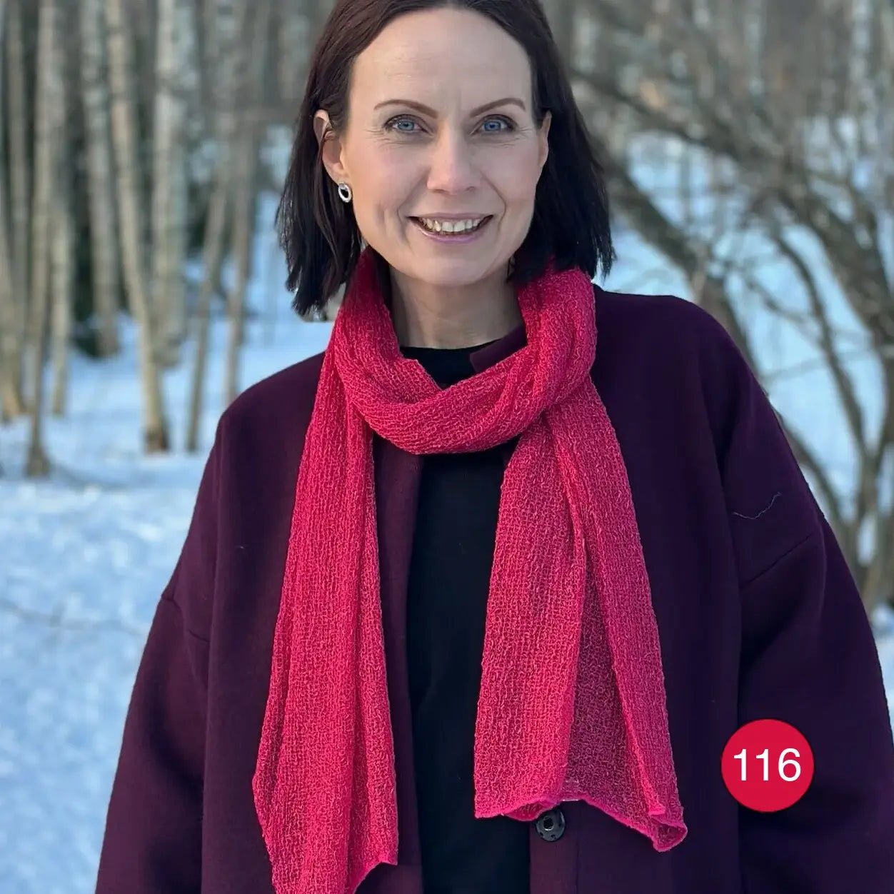 Kvinna utomhus med en Almira sjal i azaleröd ton virad runt halsen över en mörkblå tröja; den finstickade sjalen lyser hallonröd mot det mörka. Ulla Jacobsson Kläder