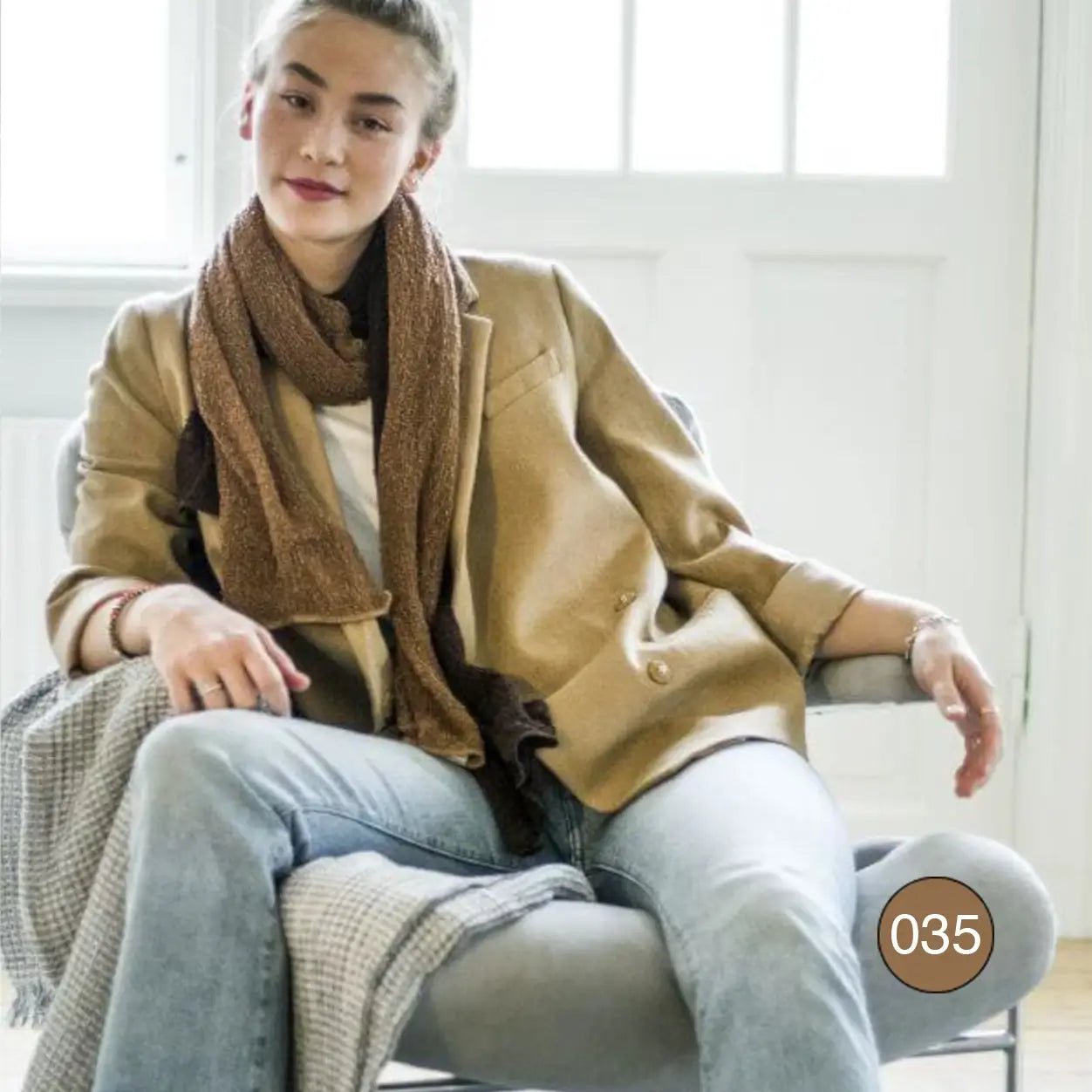Kvinna sittande i en beige kappa och jeans, med en Almira sjal i bärnsten löst lindad runt halsen; det varma stickade garnet ramar in ansiktet. Ulla Jacobsson Kläder
