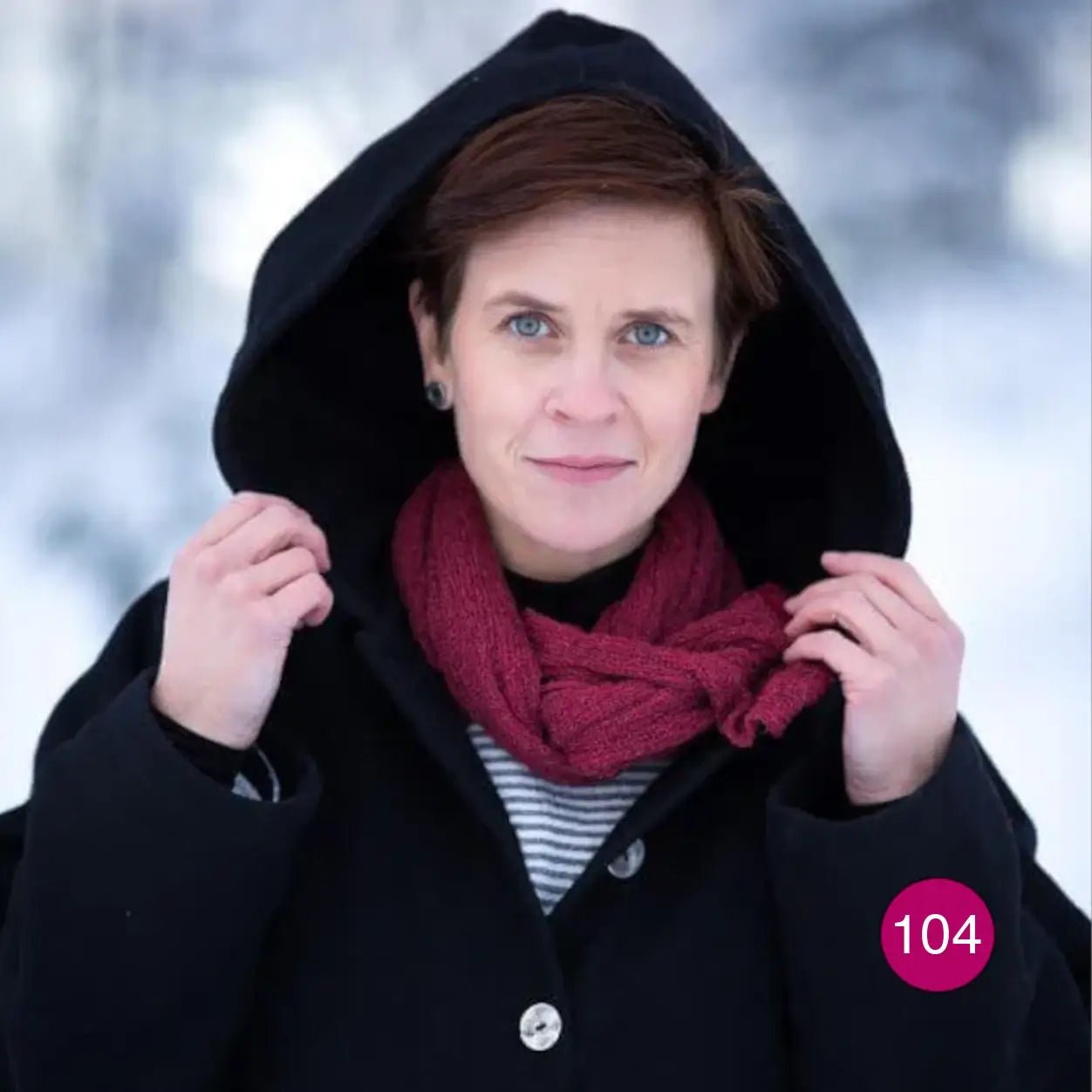 Kvinna med uppfälld huva på en mörk kappa, med en Almira sjal i fuchsia virad runt halsen; det starkt rosa stickade garnet lyser mot det mörka tyget. Ulla Jacobsson Kläder