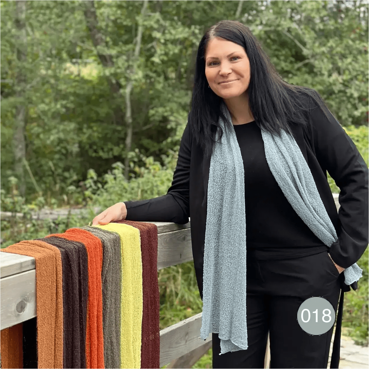 Kvinna vid ett räcke där flera färgglada sjalar hänger på rad, själv klädd i en gråblå Almira sjal som löper rakt ned över en mörk tröja. Ulla Jacobsson Kläder