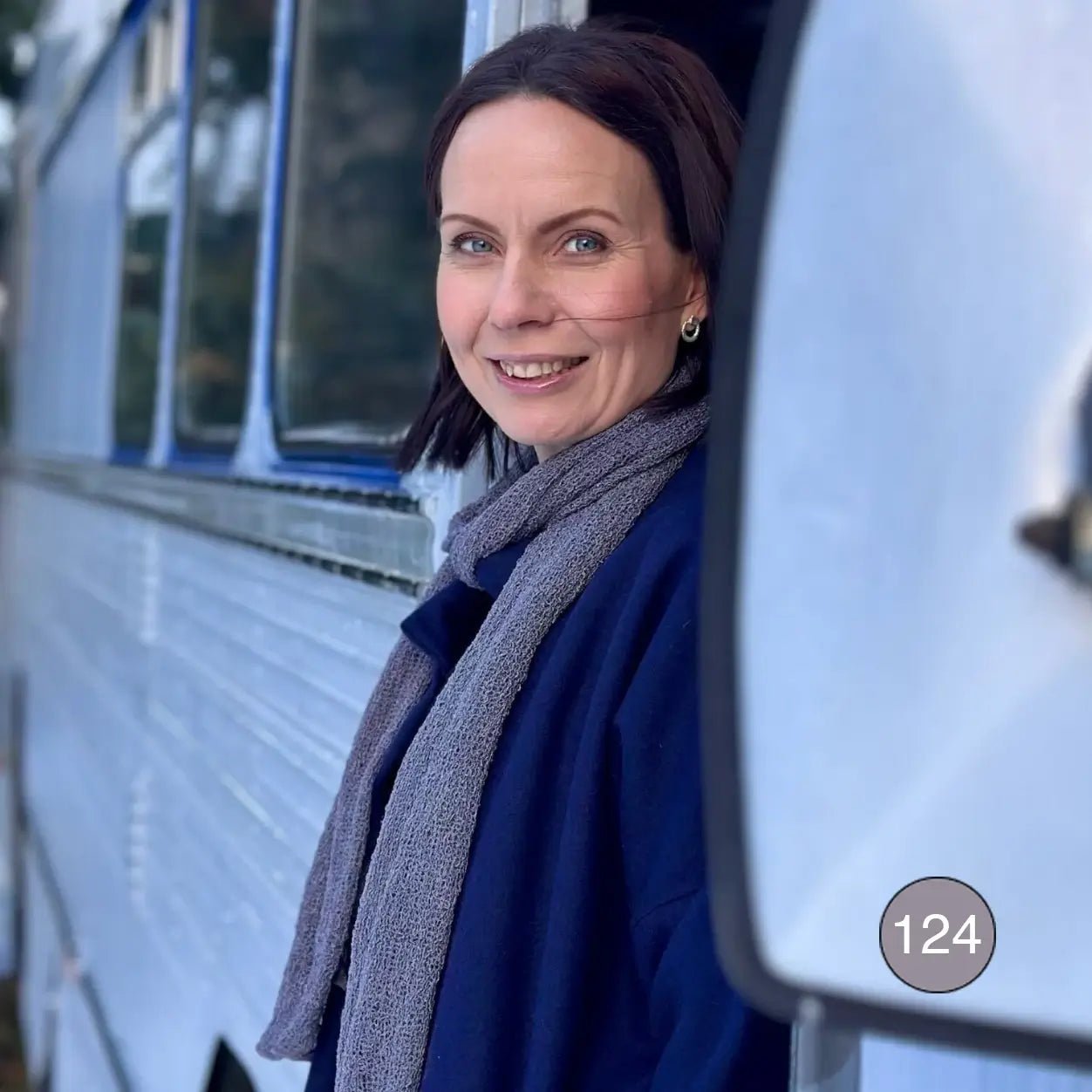 Mörkhårig kvinna utomhus vid en husvägg, med en Almira sjal i grålila lagd runt halsen; det dämpade lila garnet faller mjukt i två längder framtill. Ulla Jacobsson Kläder