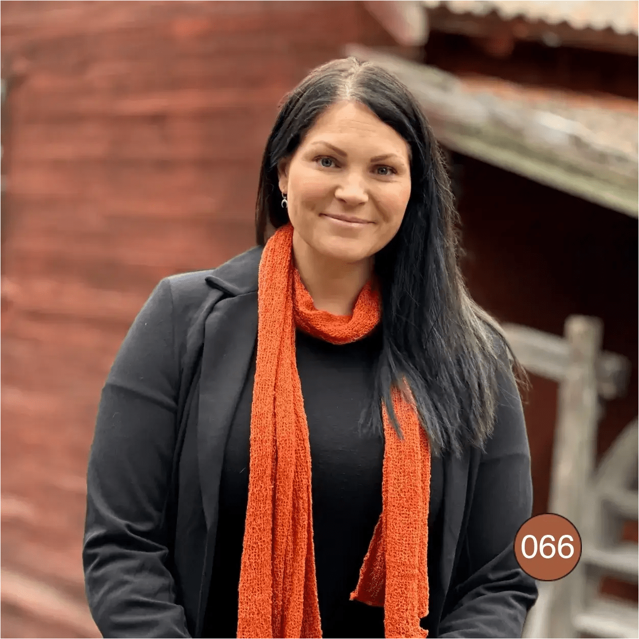 Mörkhårig kvinna utomhus framför en röd lada, med en Almira sjal i kopparröd ton virad runt halsen över en svart tröja; den långa sjalen faller varmt orangeröd nedför framsidan. Ulla Jacobsson Kläder
