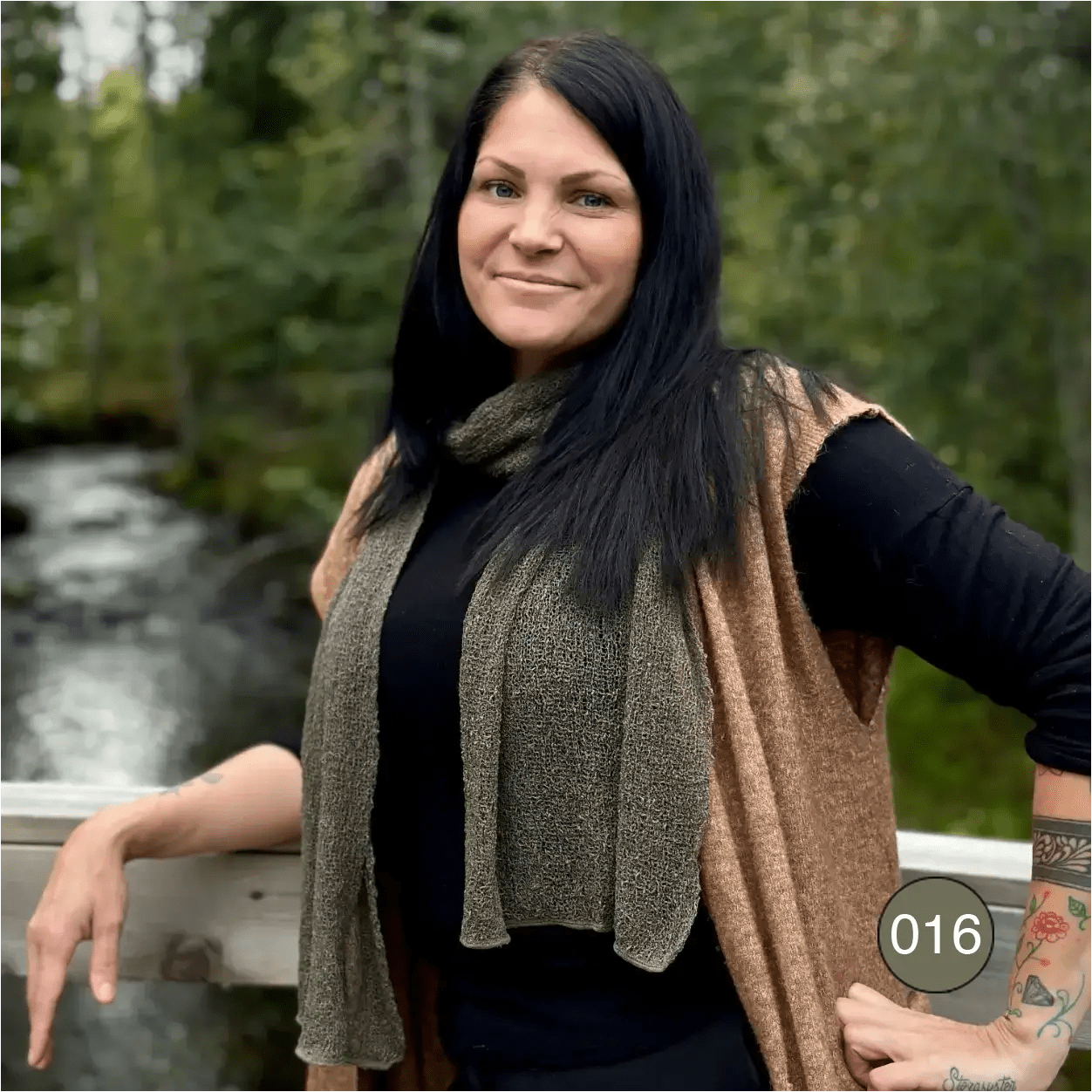 Mörkhårig kvinna med händerna i sidan bär en Almira sjal i lavgrön ton som hänger i två raka längder över en svart tröja. Ulla Jacobsson Kläder