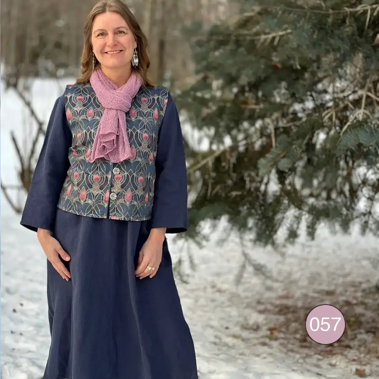 Kvinna i mönstrad jacka och lång kjol med en Almira sjal i lingon och mjölk löst draperad runt halsen; det rosa- och vitspräckliga garnet syns i helfigur. Ulla Jacobsson Kläder