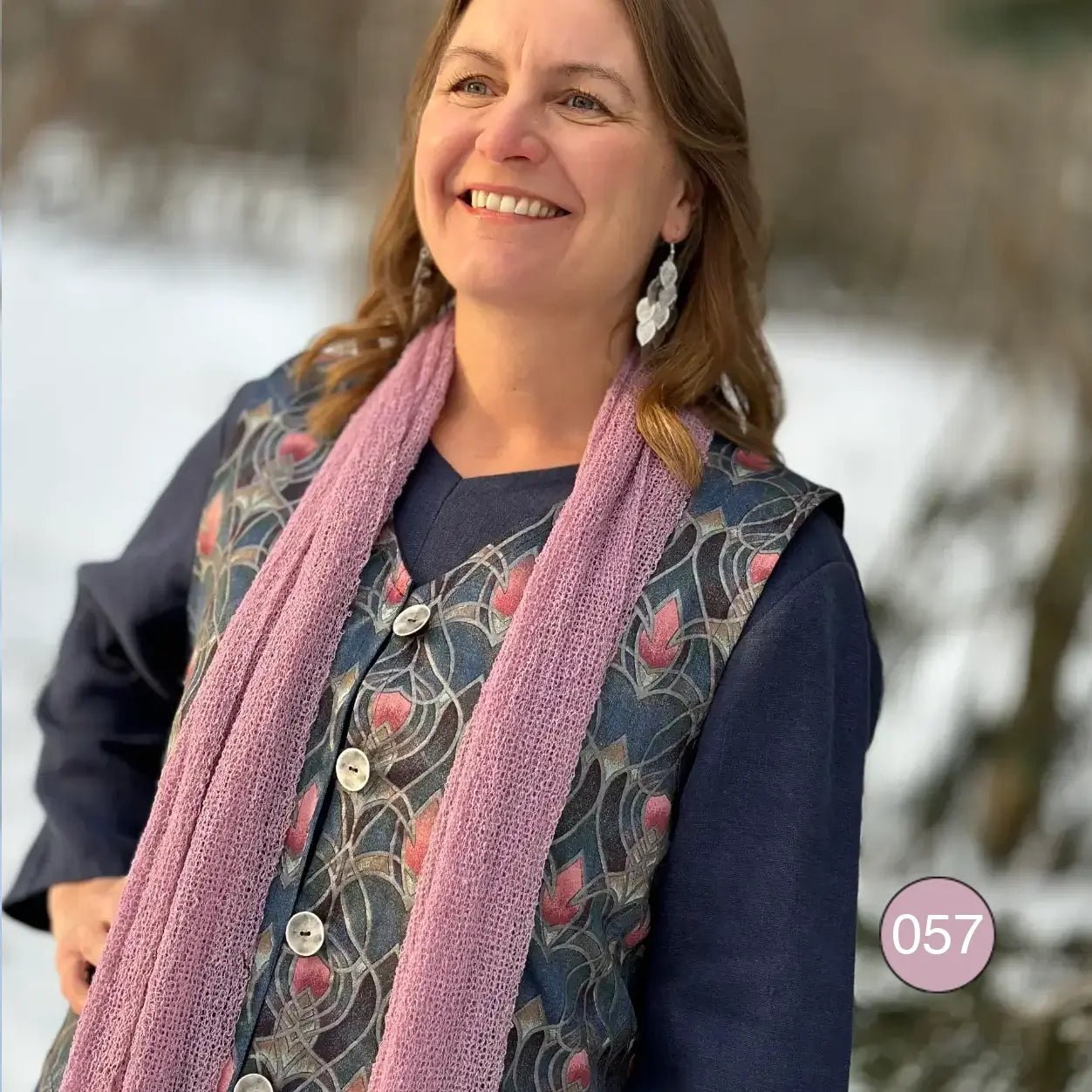 Leende kvinna i mörk topp med en Almira sjal i lingon och mjölk kring halsen; det rosa- och vitmelerade garnet faller mjukt nedför axlarna. Ulla Jacobsson Kläder